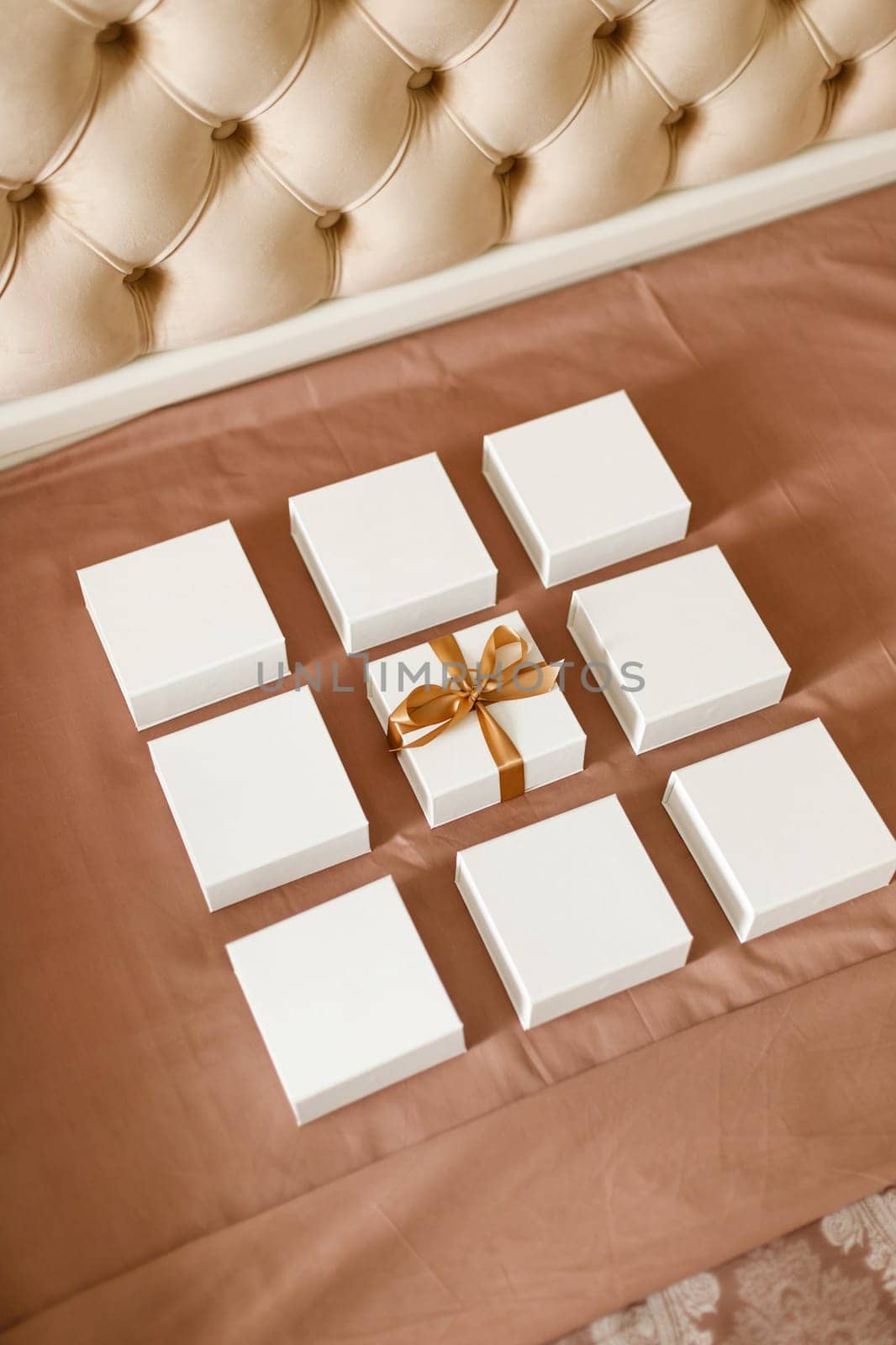 Presents for holidays. Set of small minimalist gift boxes for jewelry spread out on luxury bed. Close up of nine white square boxes one of which is tied with brown ribbon.