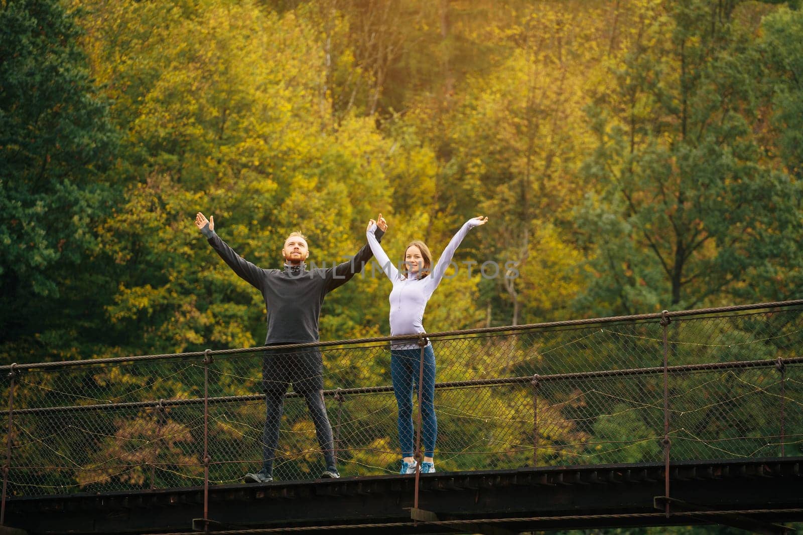 Athletic man and woman warm up before charging while walking in the mountains. Couple standing on a suspension bridge on a background of forest. Concept of tourist active life and adventure.
