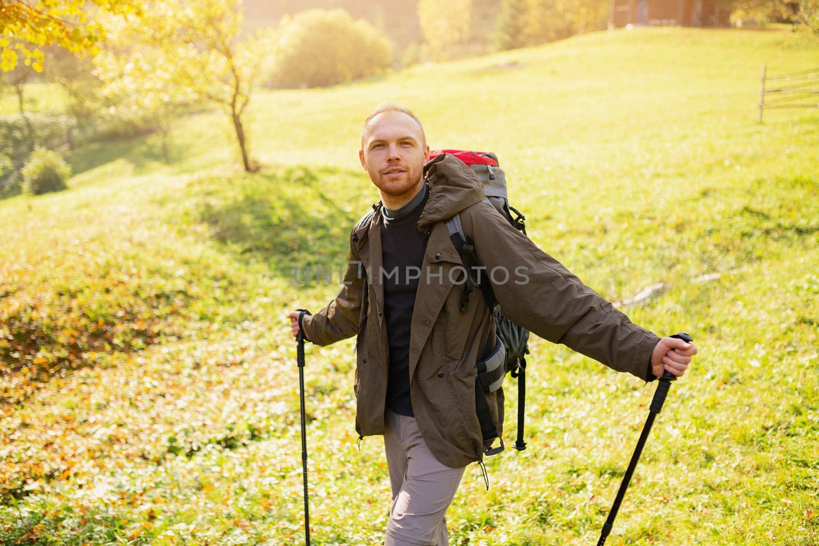 Male hiker with hiking equipment stands in the middle of a mountain village on a hill and looks at the camera. Happy guy with a backpack and trekking sticks explores the sunny mountains. Wanderlust.