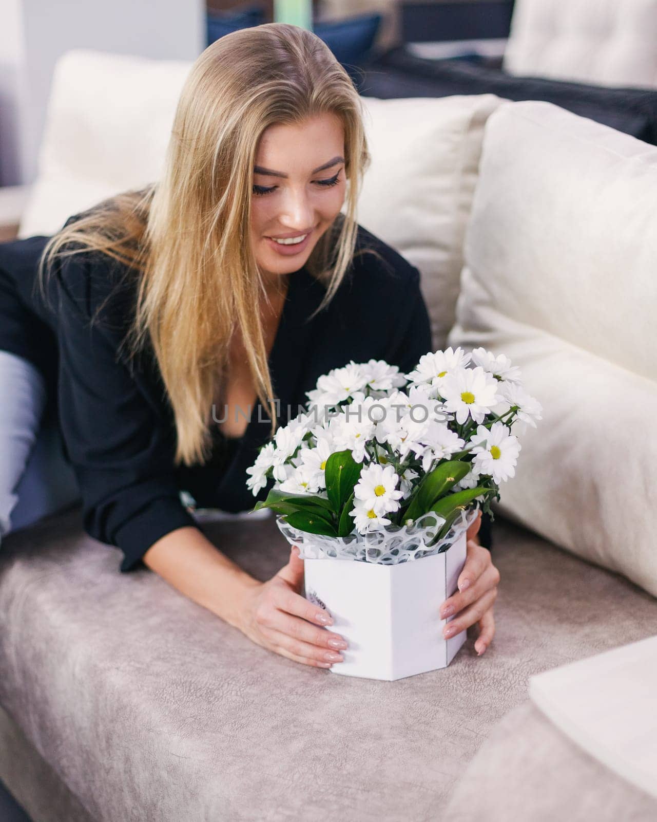 Excited and happy young woman rejoices and enjoys flowers given to her. Beautiful elegant caucasian young blonde woman sitting on gray comfortable sofa with bouquet of flowers standing in gift box.