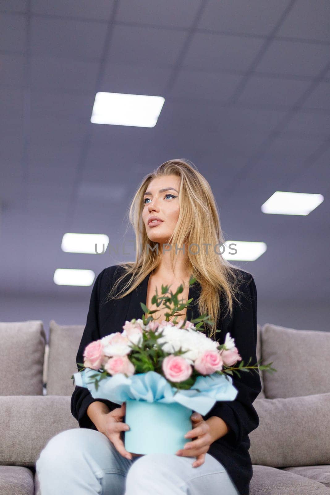 Portrait of attractive beautiful and stylish young woman with bouquet of flowers in her hands. Woman sitting on sofa and holding cardboard gift box with flowers and looking away. Bottom view.