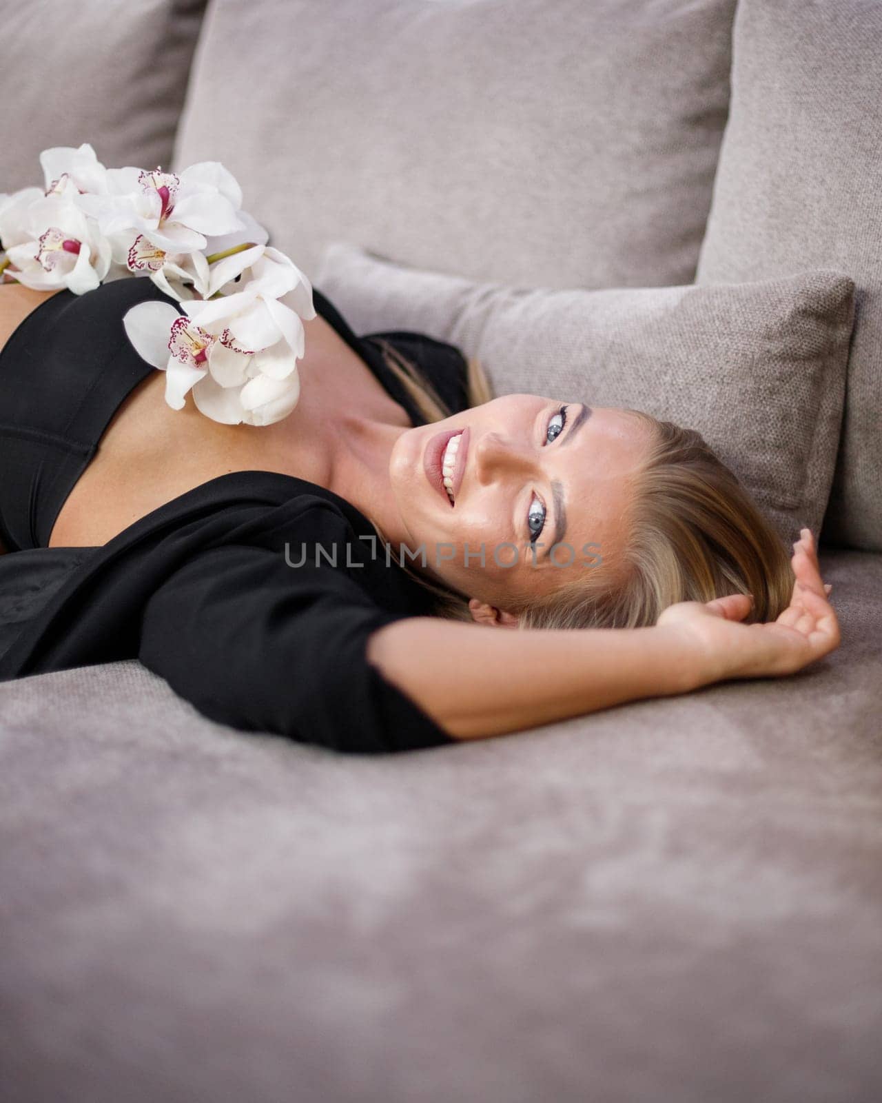 Portrait of seductive woman lying on sofa with orchid branch. Spectacular blonde woman in black top and jacket lies with flowers on her chest and looks at camera. Concept of femininity.