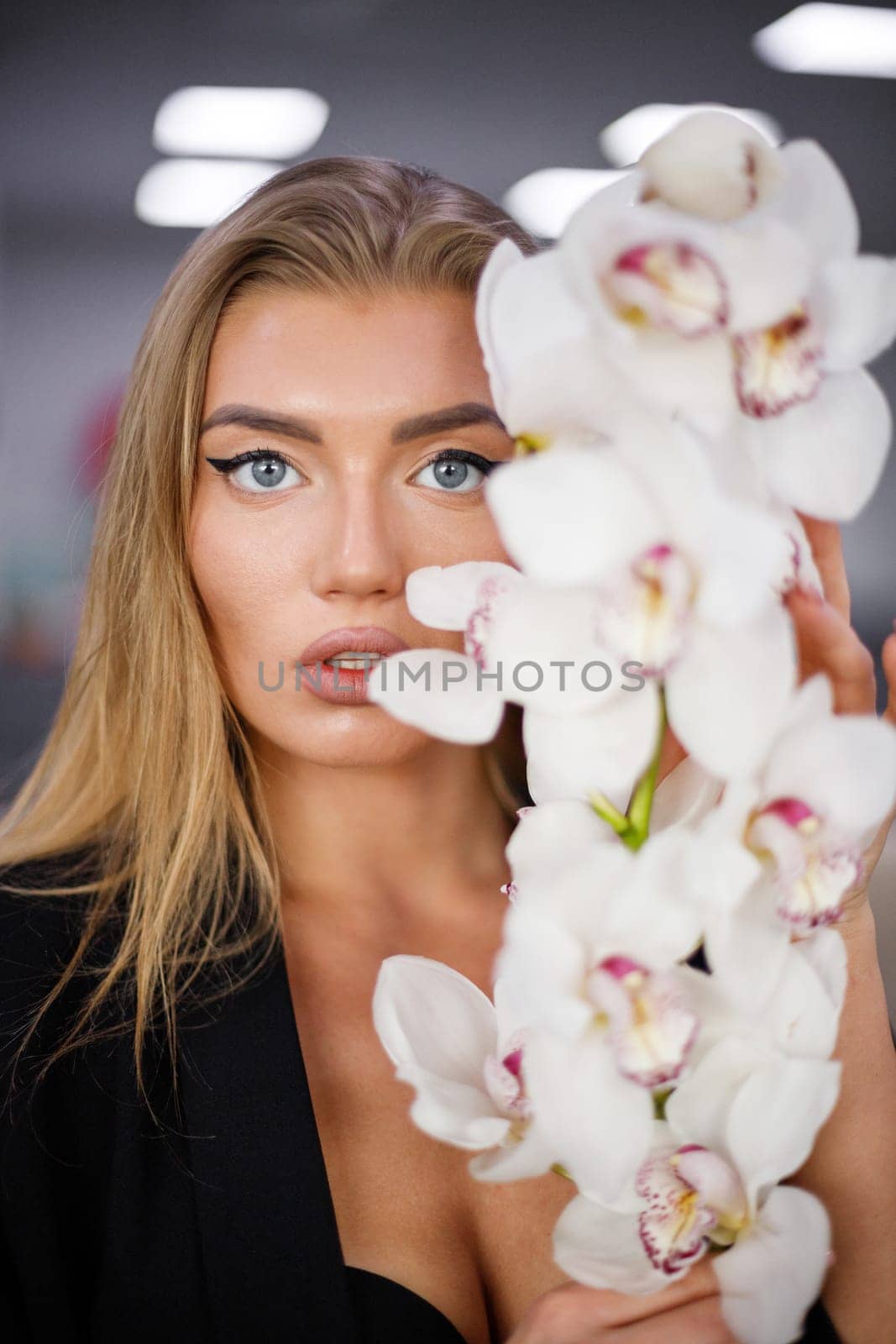 Portrait of fashionable beautiful young woman with orchid branch near face. Close up of stylish caucasian blonde woman with makeup wearing black jacket posing with fresh orchid flower. Selective focus