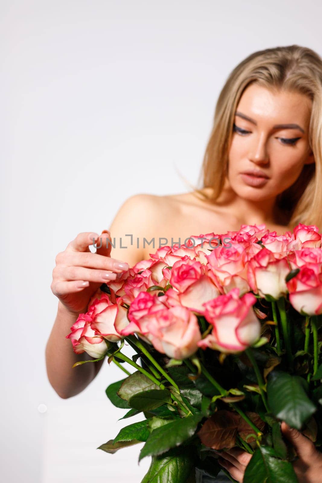 Portrait of naked young woman with large bouquet of roses covering her body. Spectacular blonde woman with makeup looks at large number of pink roses standing on white background. Copy space.