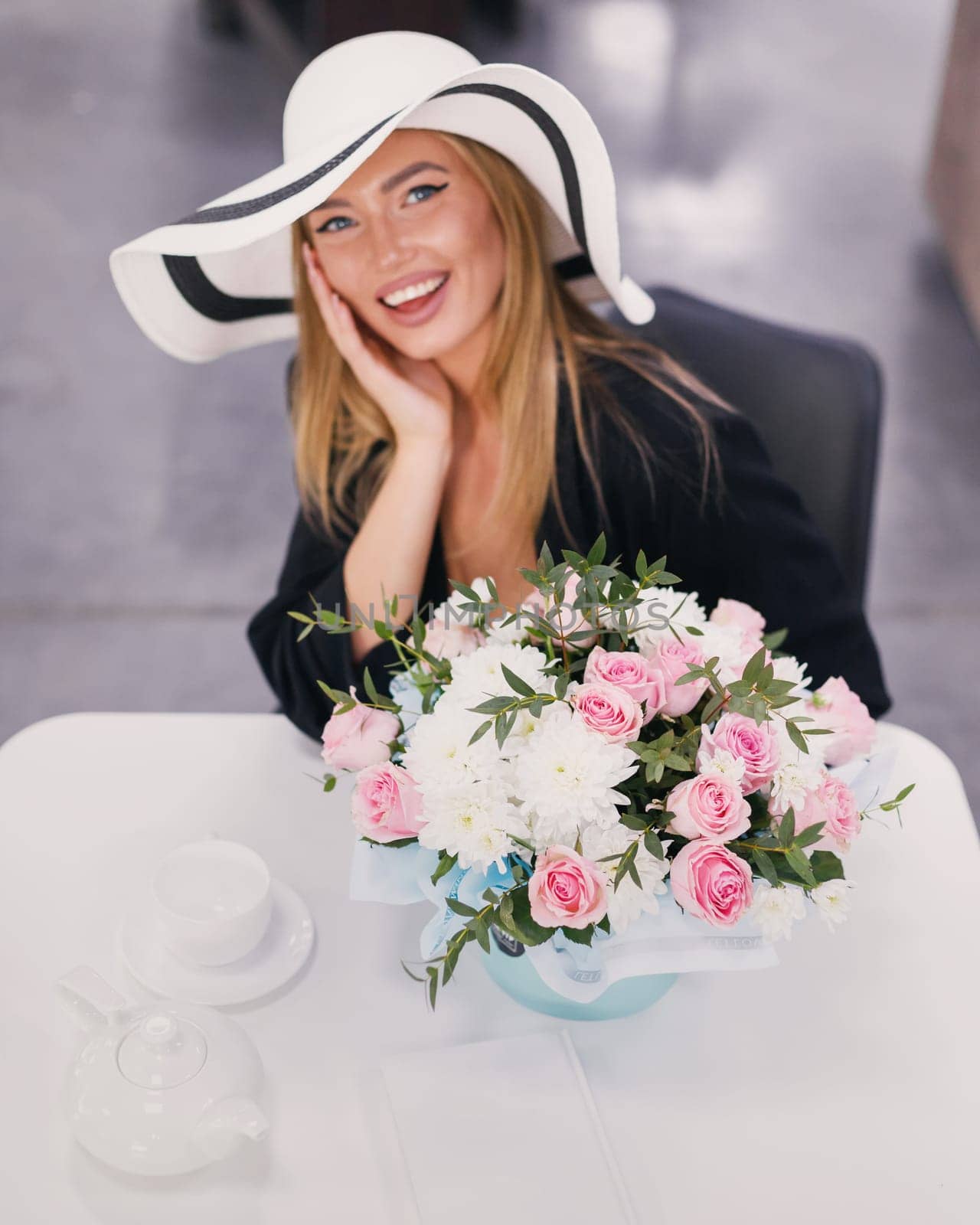 Fashion model portrait. Stylish beautiful young woman resting in restaurant sitting near bouquet of flowers. Woman with make-up dressed in white hat sits at table with teapot, cup and box with flowers.