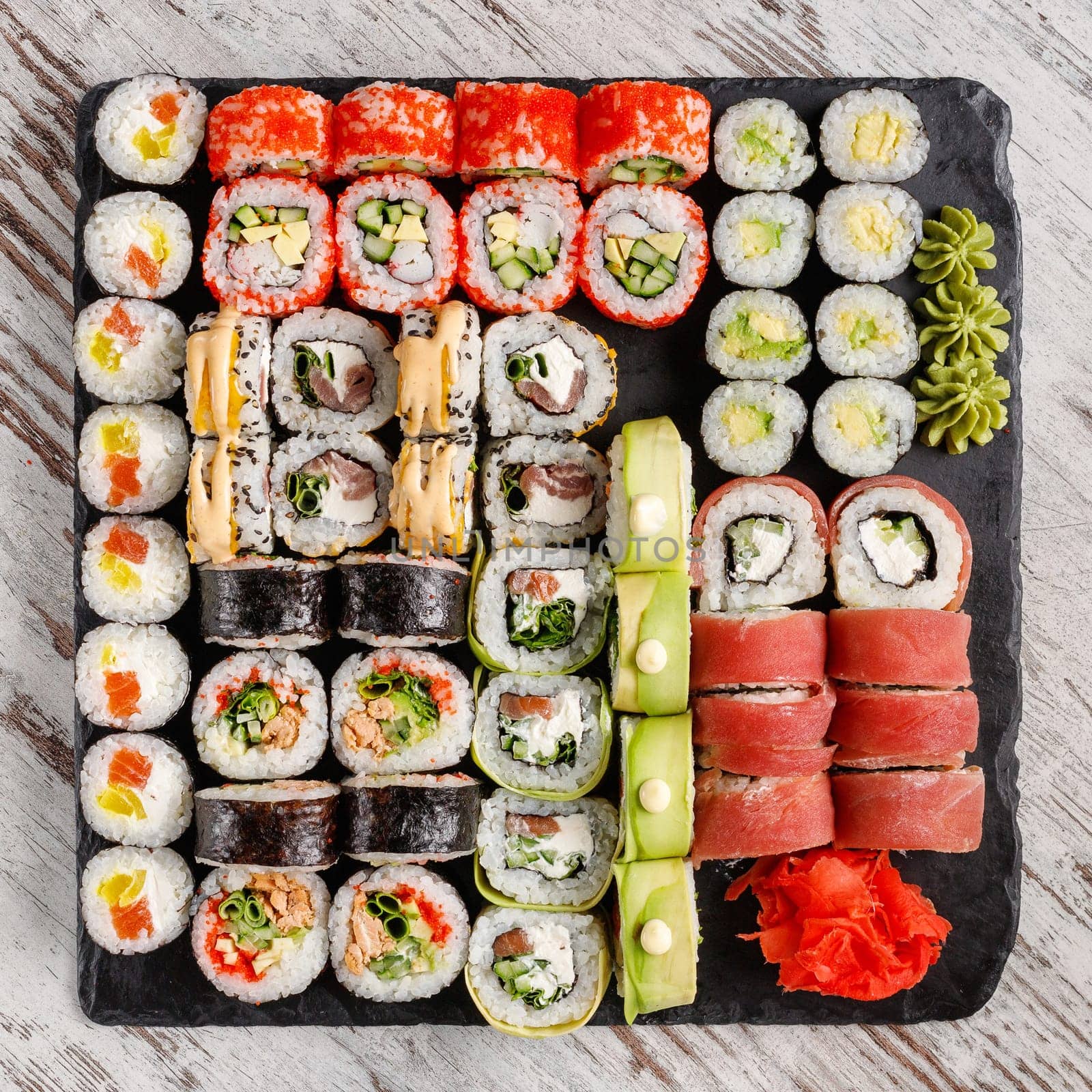 Large assorted variety of sushi rolls served on black metal slate on wooden table. Top view of sushi set consisting of sushi rolls with different fillings, makki sushi and wasabi and ginger on edges.