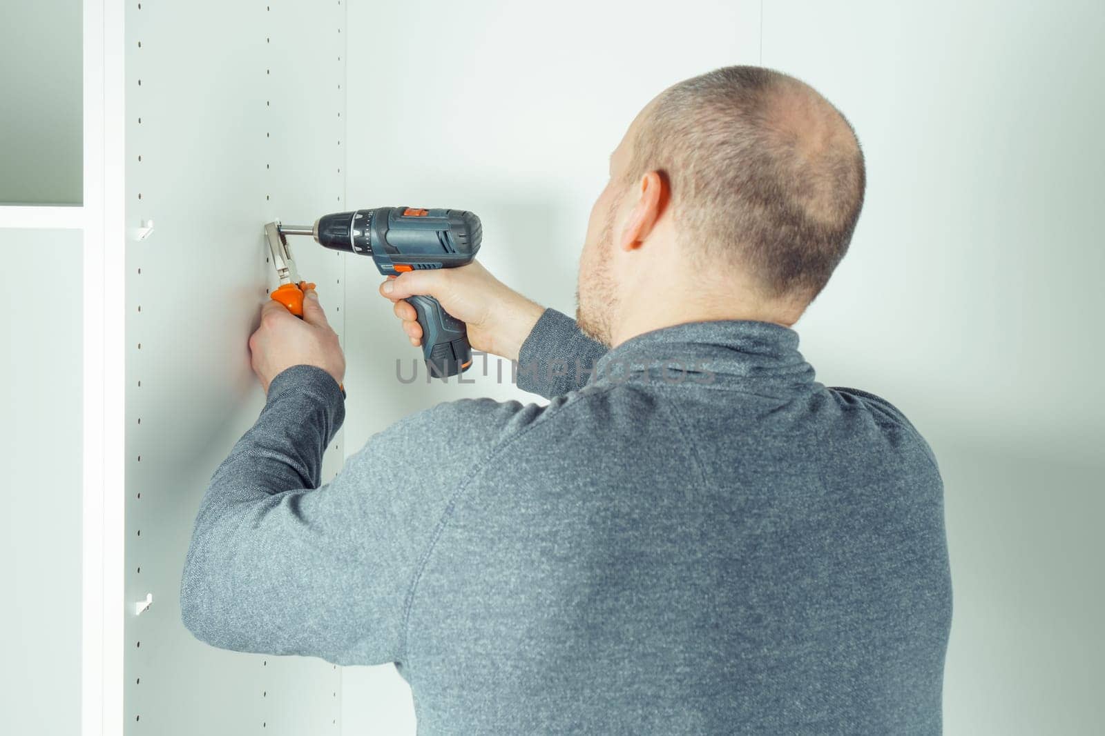 Male screw in to insert plastic support into one of series of holes to fix white shelf, assembling dresser or wardrobe. Holds screwdriver with pliers. Self-assembly of furniture, DIY, renovation