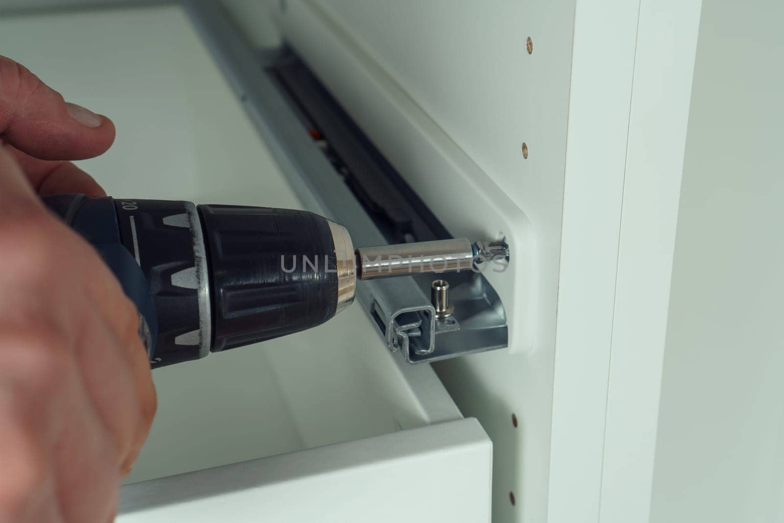 Male hands hold cordless electric screwdriver, screw to fix rail, assembling drawer of white dresser or wardrobe. Self-assembly of furniture at home, DIY, furniture repair and rearrangement