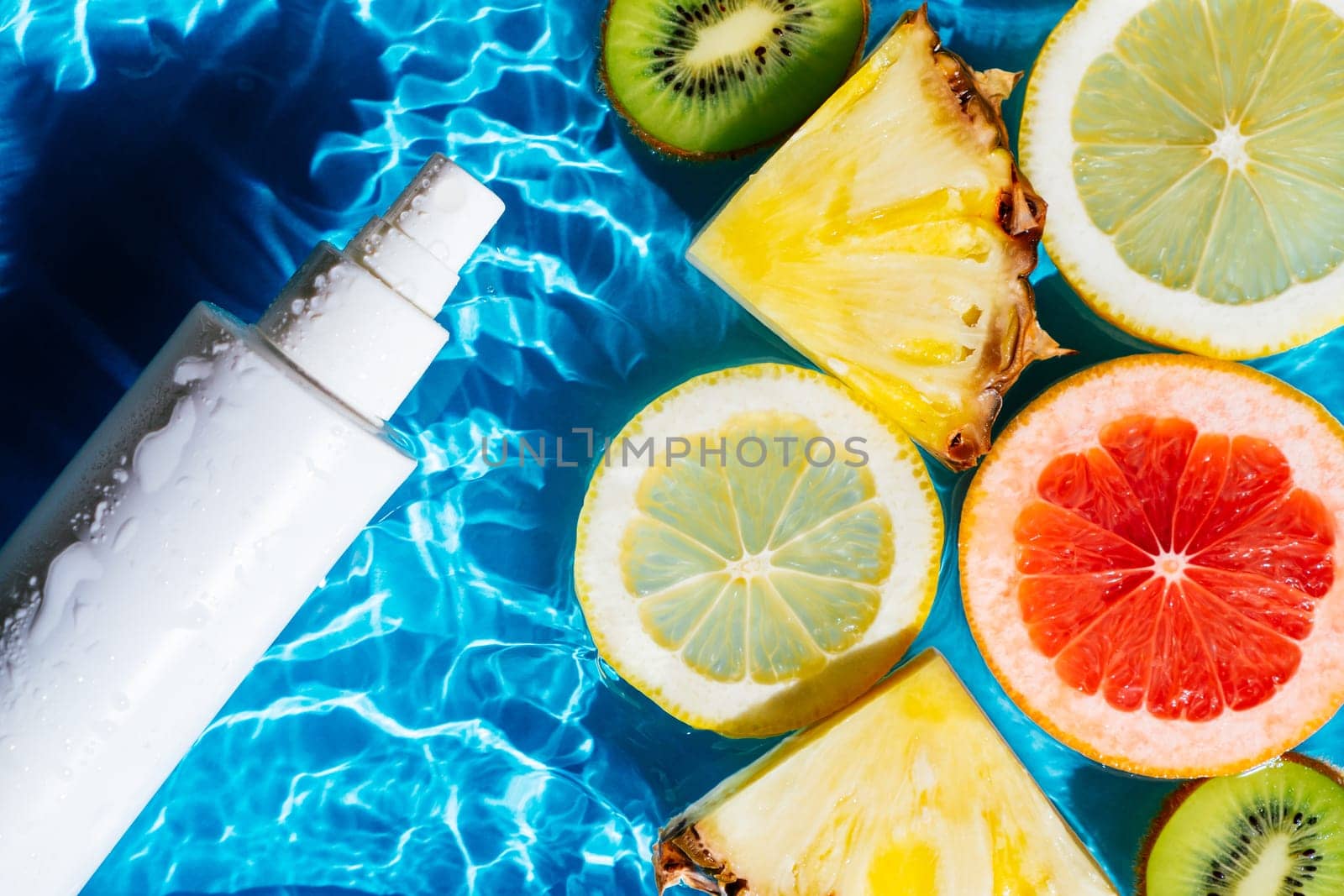 White beauty product sunscreen cream vial sprayer in transparent water with flecks and waves, fruit citrus lemon, kiwi, orange, pineapple, grapefruit. Body skin care protection beach concept. Flat lay