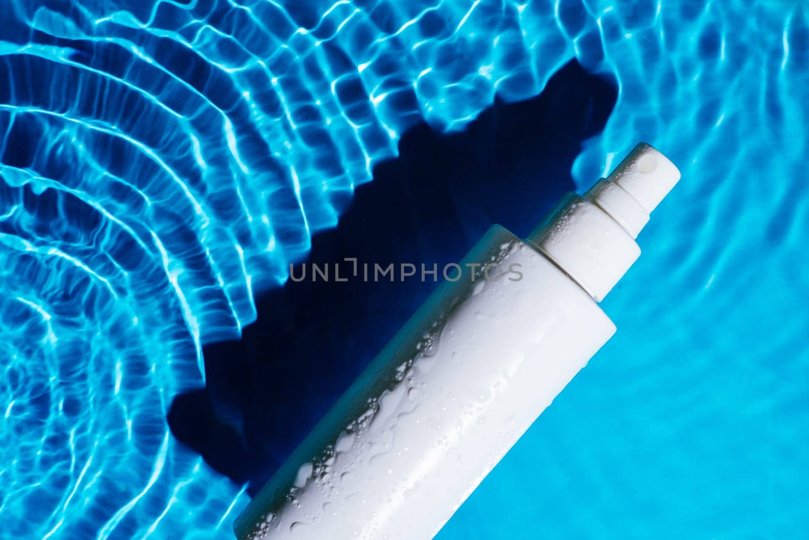 White cosmetic product moisture balm atomizer with pump vial sprayer in transparent fresh blue water with flecks, waves, shade. Body skin care protection in summer beach vacation concept. Flat lay.
