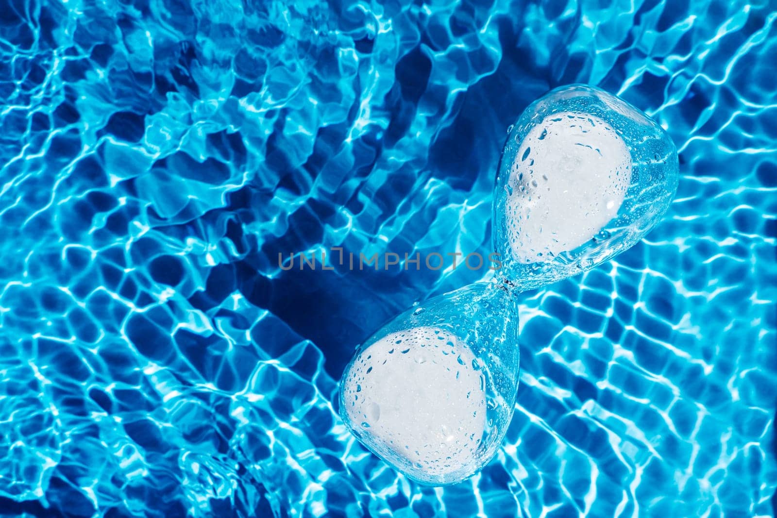 Blue abstract glass hourglass with white sand in water reflect surface pool. Renovation of time. Skin body care, anti age retirement concept. Moisturing dehydrated process. Deadline time. Flat lay.