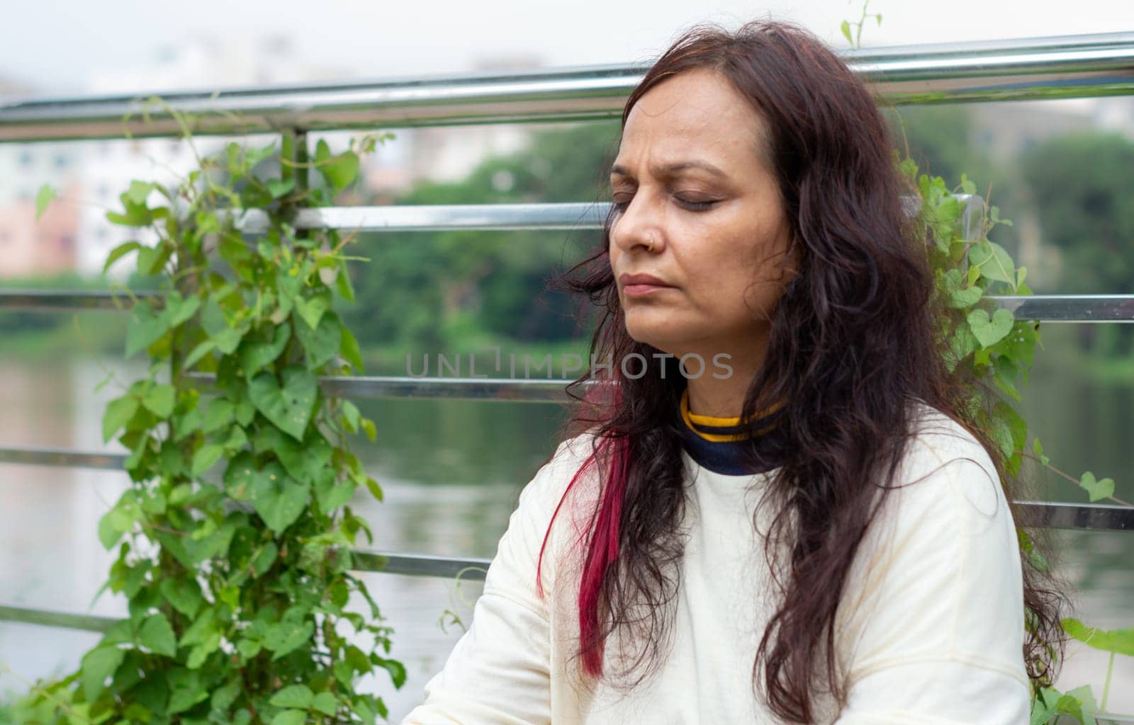 Active Mid Adult Woman in meditation sitting with eyes closed concentrate in a public park against garden leaves in the background. Front view. Head and Shoulder Shot. Close up Portrait.