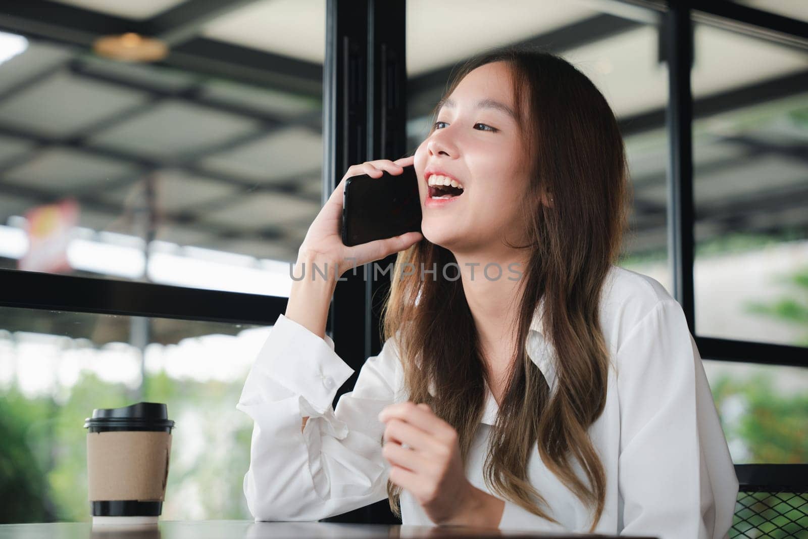 Happy female lifestyle sitting in cafe near window she cellphone on mobile phone, Asian young business woman freelance holding smartphone and coffee paper cup at coffee shop