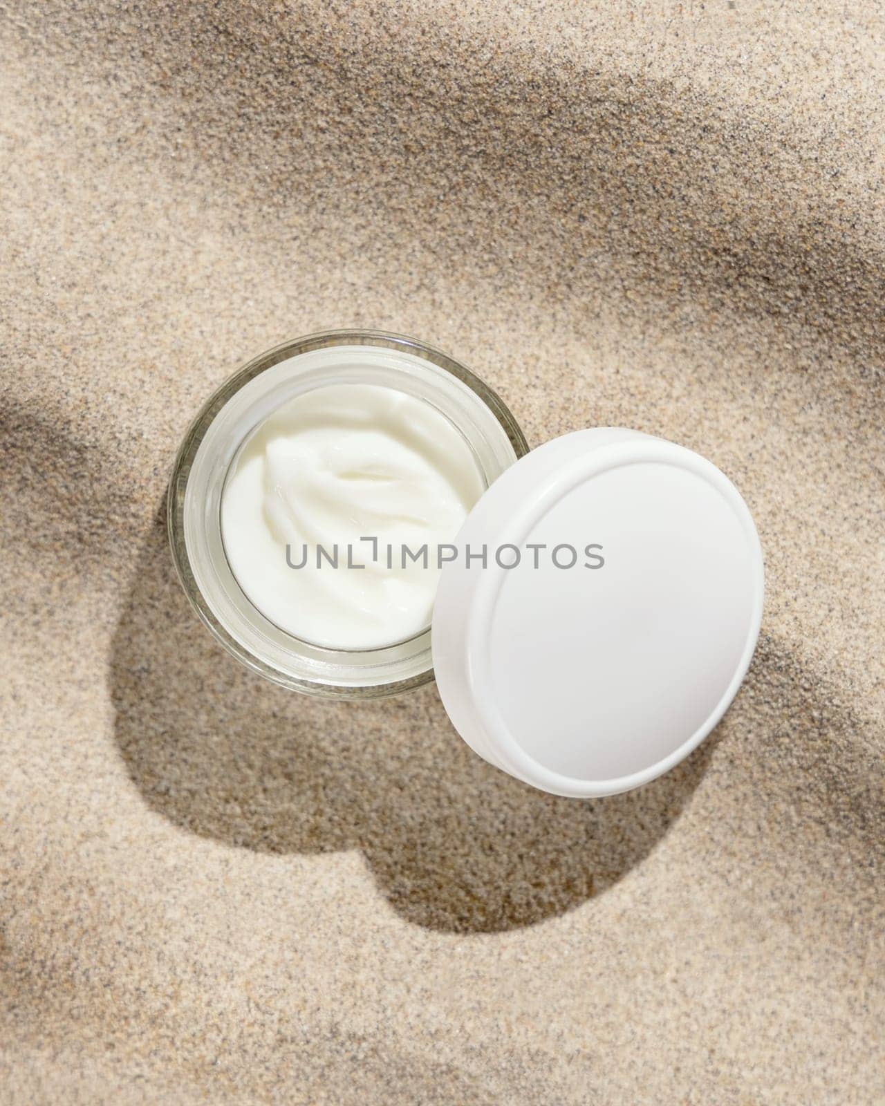 Opened cream jar with white lid on beige sand top view, hard light. Package mockup. Natural skincare product. Facial cosmetics for beauty routine. Zen style, minimal aesthetic