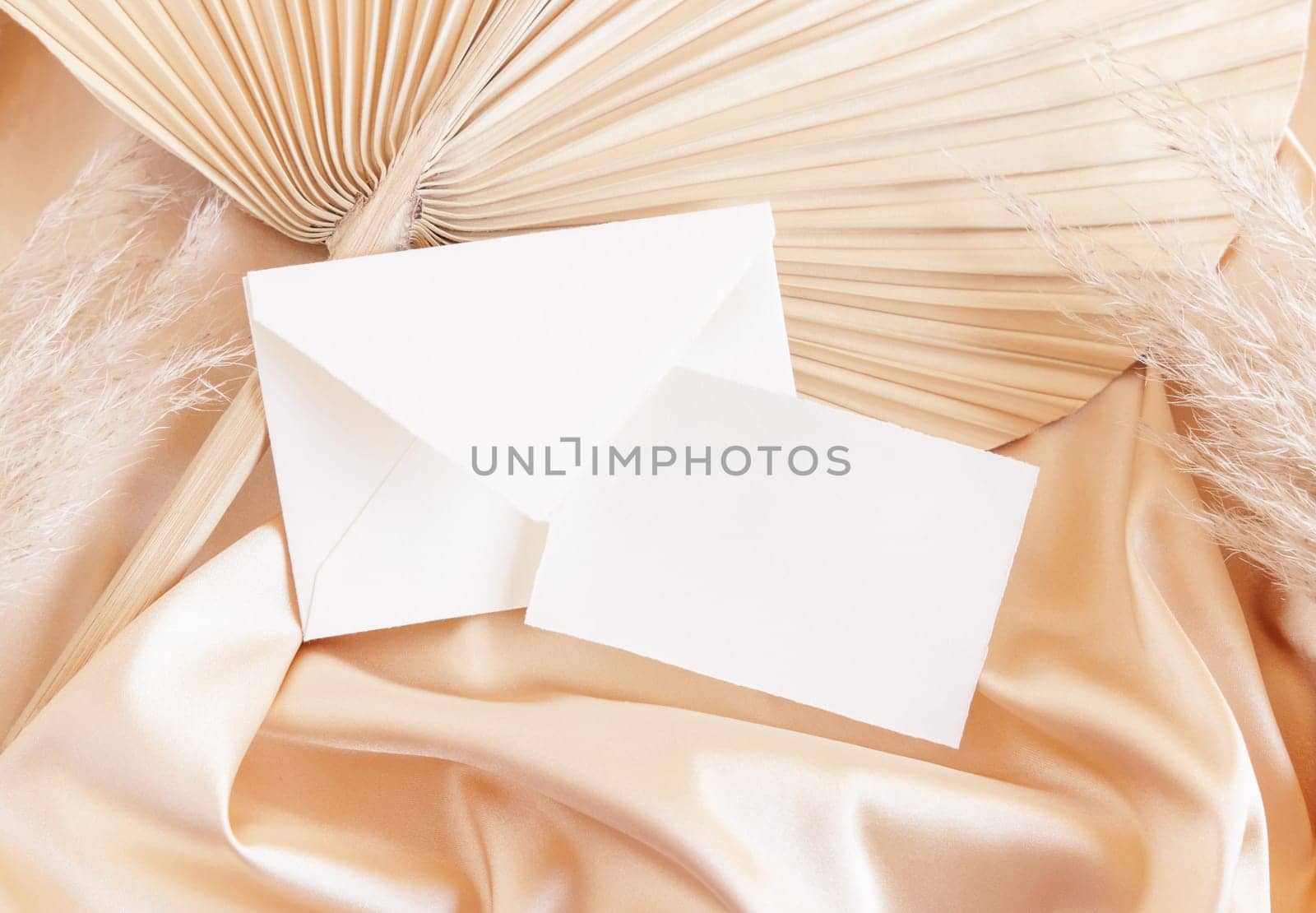 Card and envelope on beige satin fabric and dried palm leaf close up, mockup. Romantic Boho scene with horizontal paper card and envelope. Bohemian or close to nature greeting or wedding Invitation.