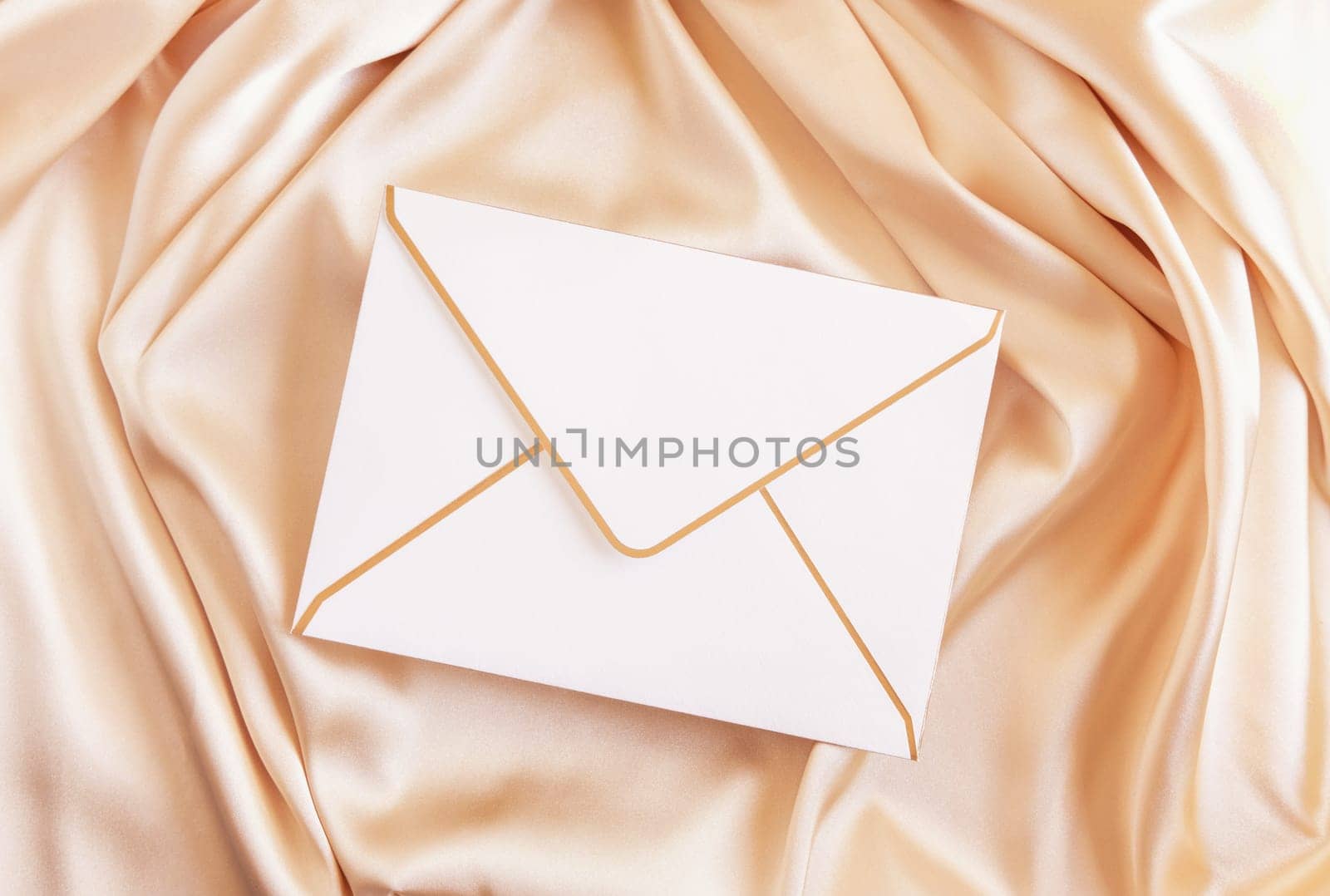 Blank envelope on beige silky fabric drapery top view, mockup. Romantic scene with white envelope, greeting or wedding Invitation.