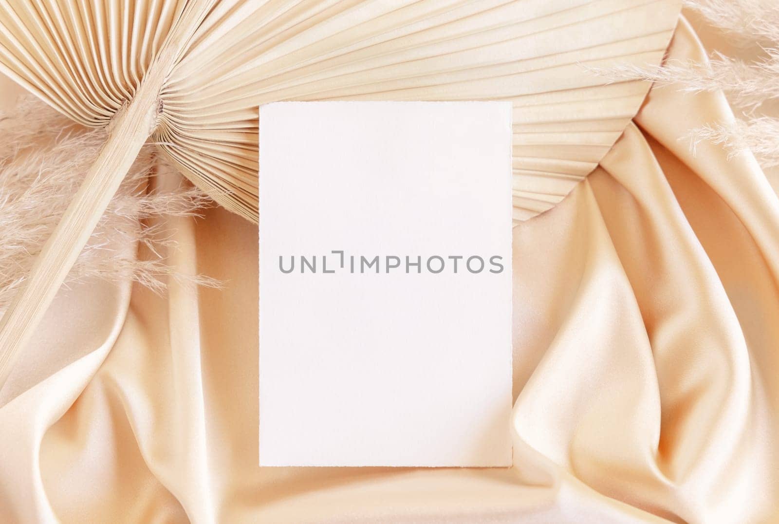 Blank card on beige satin fabric and dried palm leaf close up, mockup. Boho scene with vertical paper card. Bohemian or close to nature greeting or wedding Invitation. Romantic composition.