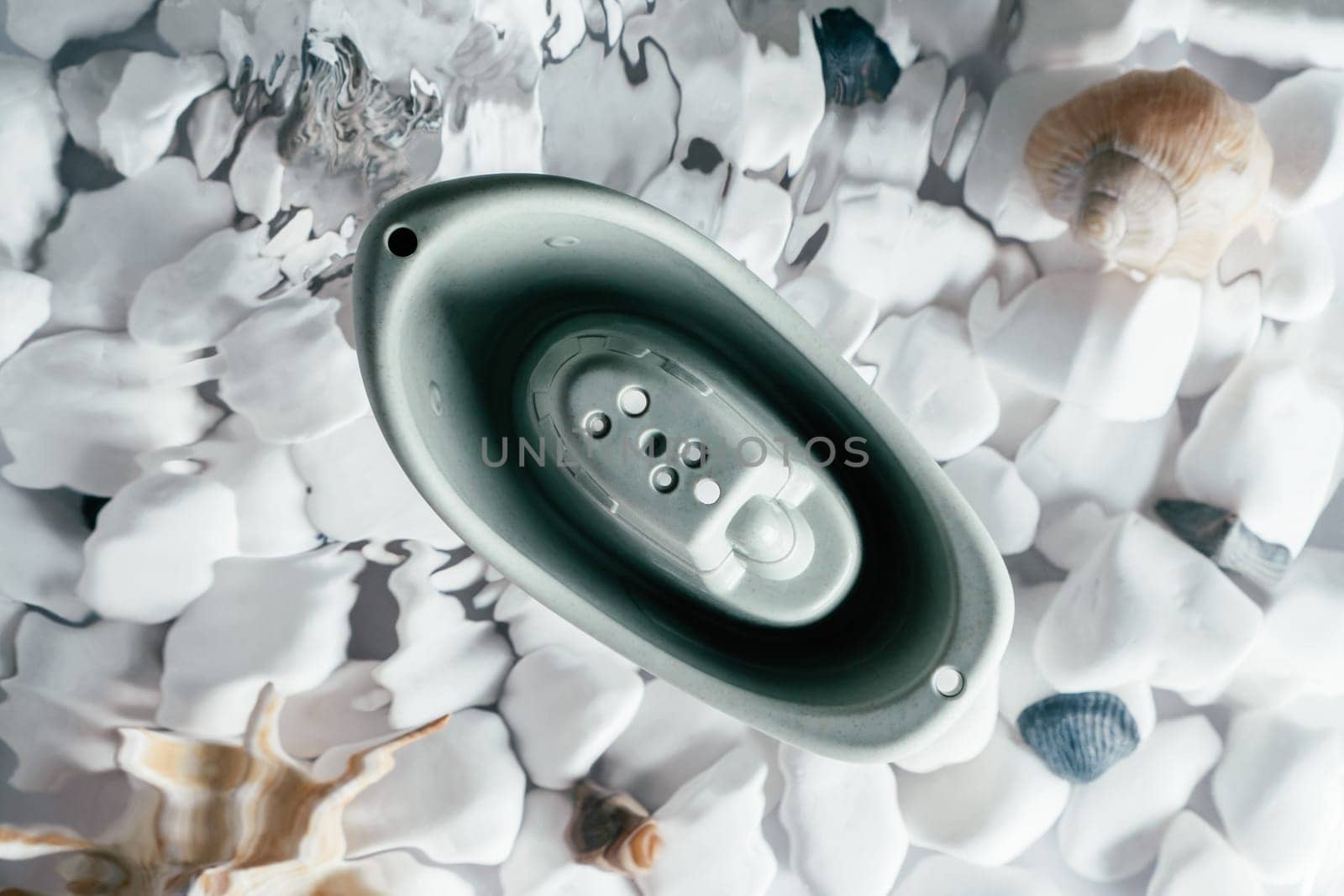 Cute grey small silicone toy boat with holes on top floating on surface of water above bottom with white stones and shell. Transparent fresh water with flecks and waves. Top view. Baby toy, water game