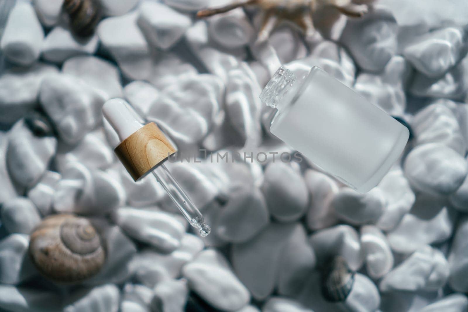 Facial pipette serum oil, essence cosmetic product in bottle on transparent fresh water with fleck and waves, bottom of white stones and shells. Organic skin body antiage care in hot climate. Top view