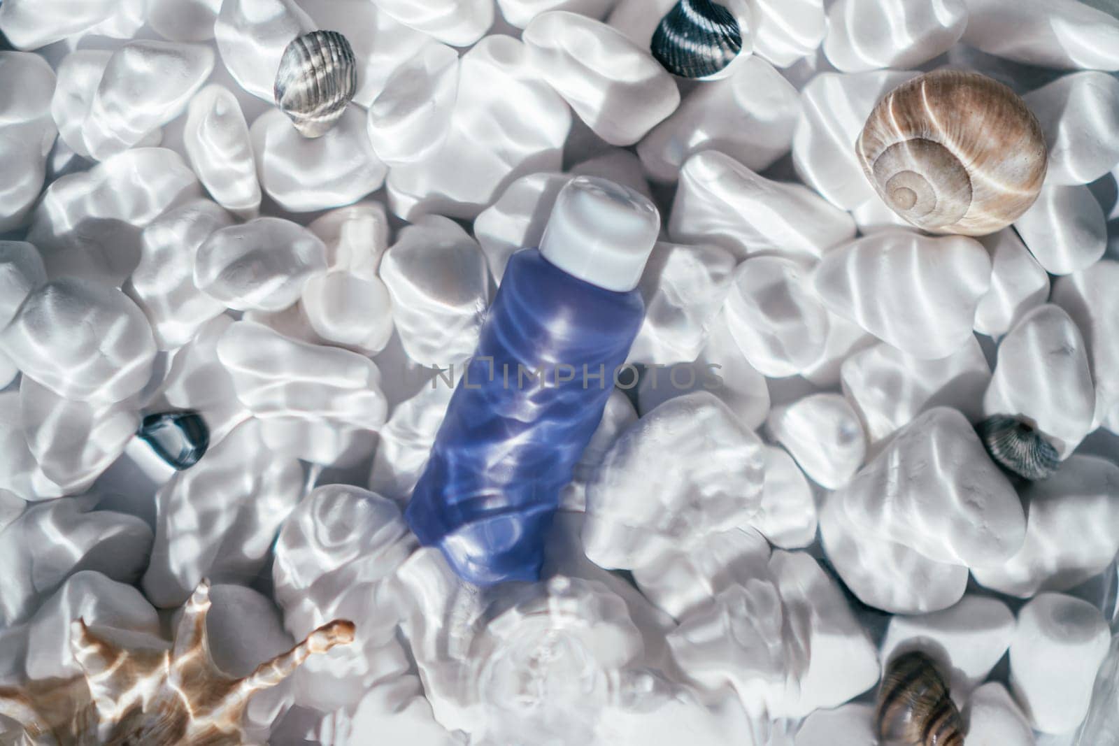 Lilac trendy bottle vial tube with white cap under transparent fresh water with flecks and waves on bottom of white stones and shells. Face and body skin care, moisturizing cosmetic. Top view