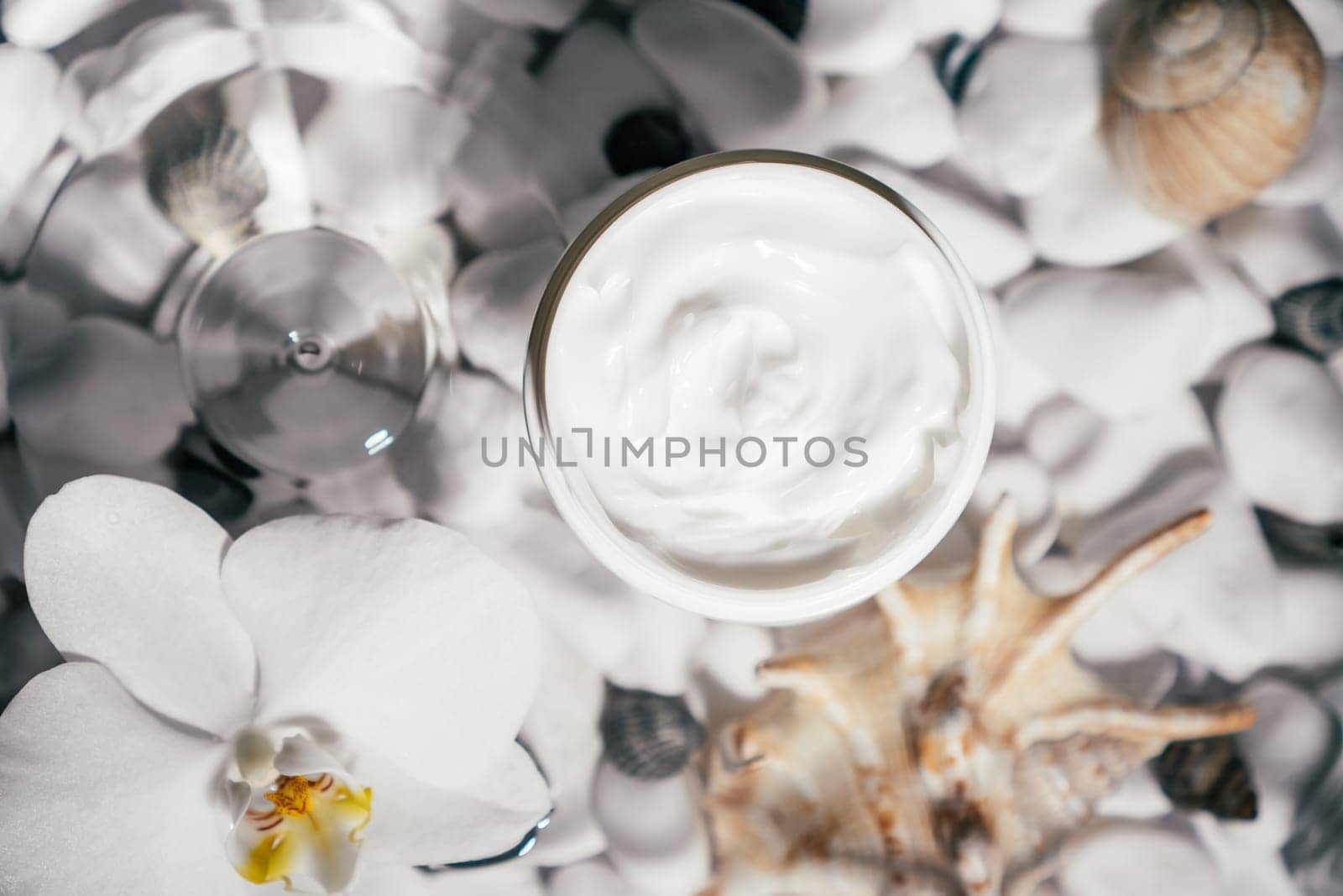 Facial jar cream milky beauty product and white orchid on transparent fresh water with flecks and waves, bottom of white stones and shells. Face and body skin care, moisturizing cosmetic. Top view