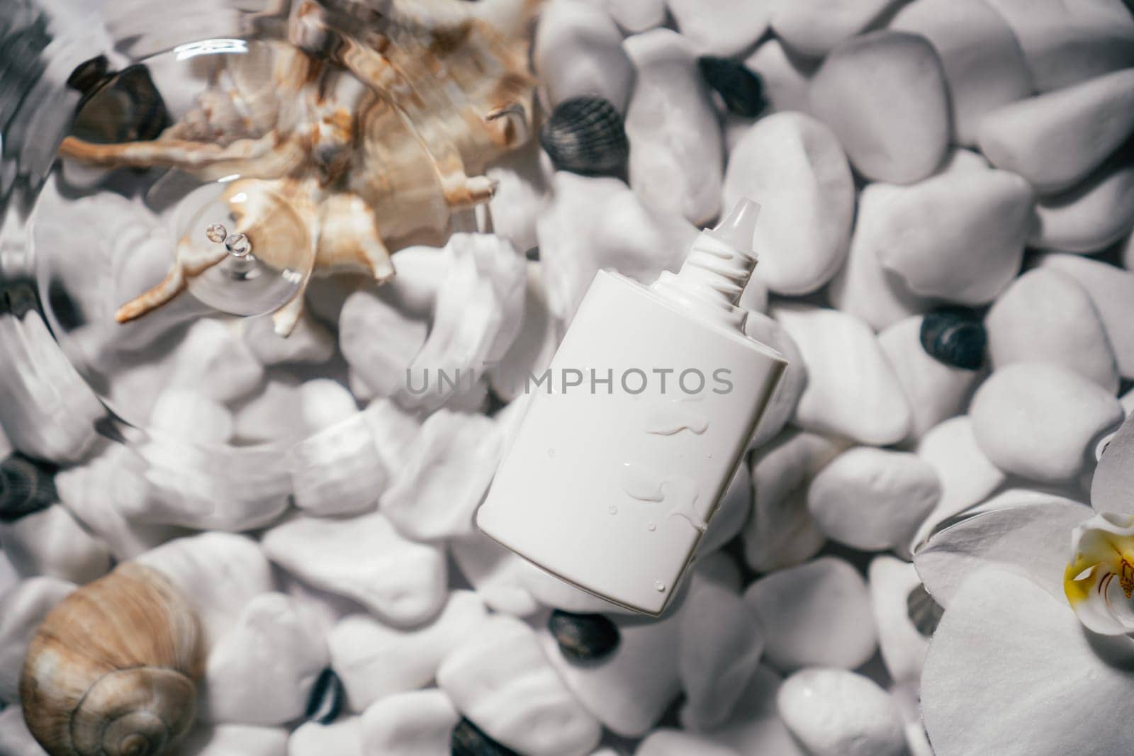 White beauty product sunscreen cream vial tube bottle on transparent fresh water with flecks and waves, bottom of white stones and shells. Face and body skin care, moisturizing cosmetic. Top view