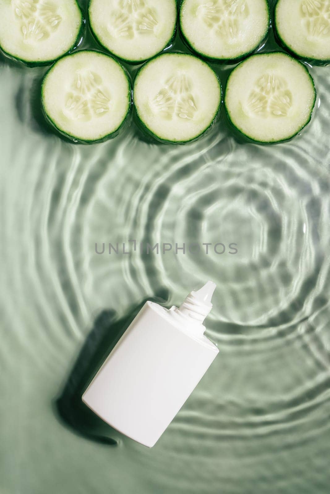 White beauty product cream vial tube bottle with rows of cucumber slices on surface of rippled transparent green water with fleck, wave, shadow. Concentric expanding circles from fallen drop. Vertical