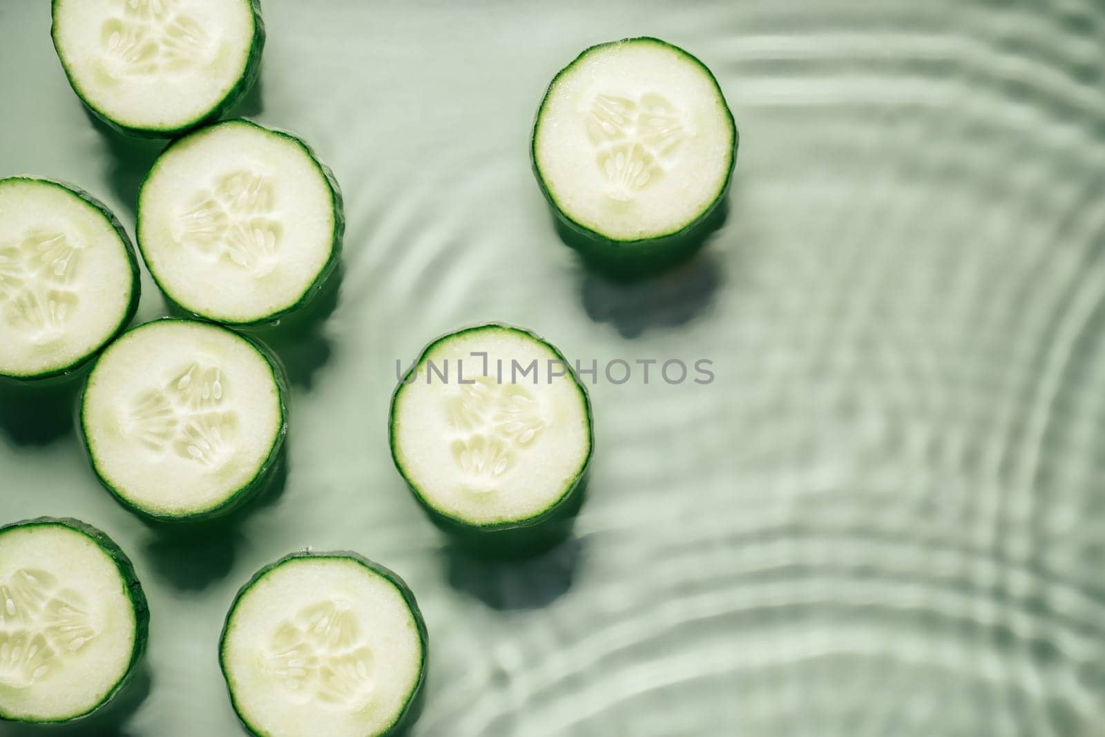 Cucumber slices lie on surface of rippled transparent fresh green exhilarant water gel with flecks, waves, shadow. Concentric expanding circles on surface from fallen drop. Health care. Copy space