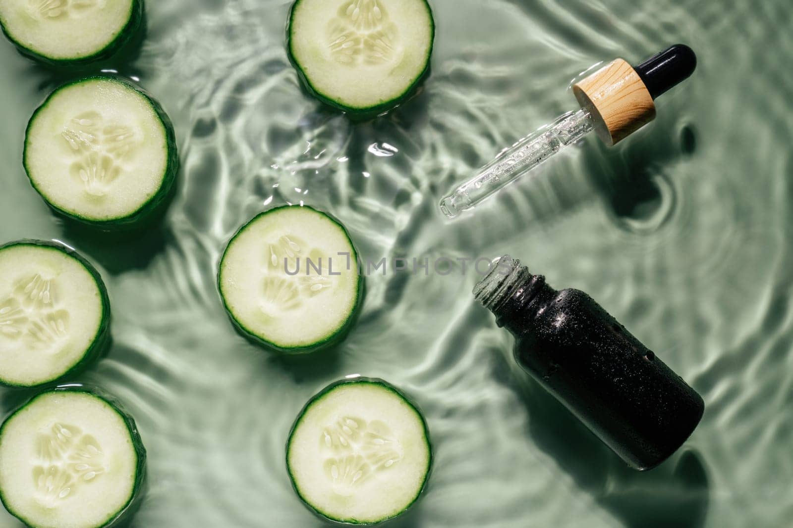 Facial pipette serum black bottle, whey beauty product near cucumber slices in water splashing fresh transparent, flecks. Flat lay, top view. Organic skin face and body moisturizing and rejuvenating
