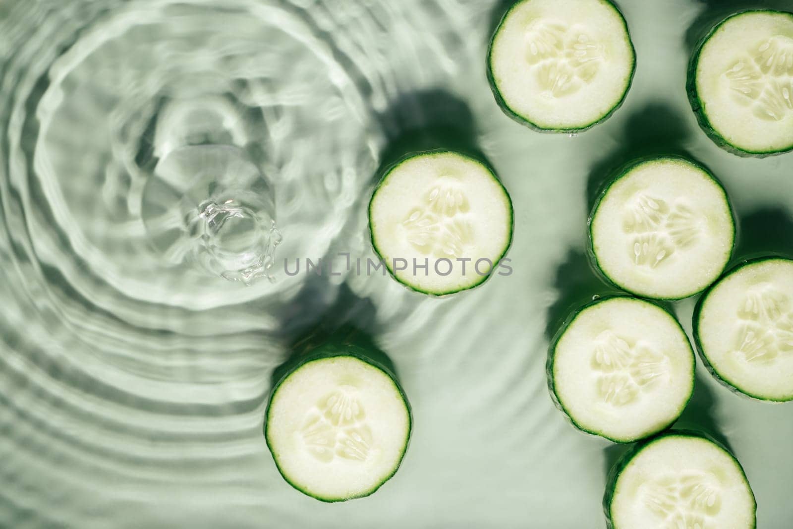 Cucumber slices lie on surface of rippled transparent fresh green exhilarant water gel with flecks, waves, shadow. Concentric expanding circles on surface from fallen drop. Health care. Copy space
