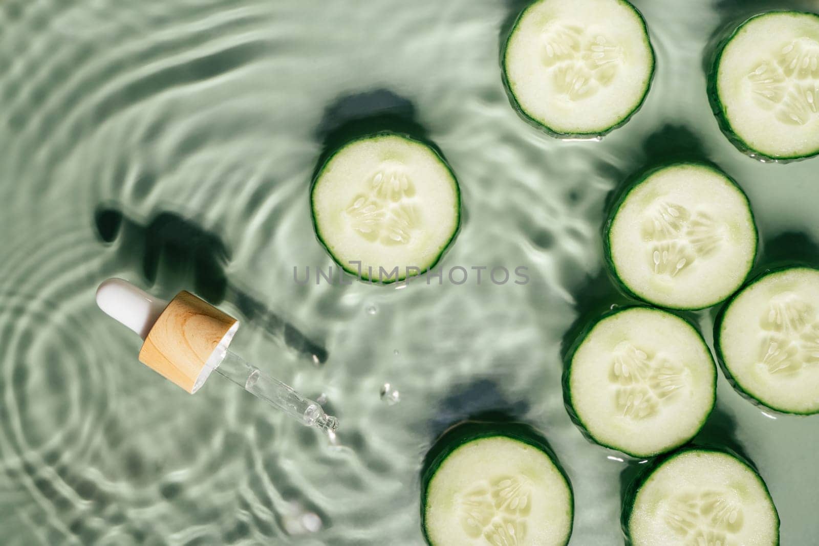 Facial pipette serum, whey beauty product near cucumber slices in water splashing fresh transparent, flecks. Flat lay, top view. Summer beach. Organic skin face and body moisturizing and rejuvenating