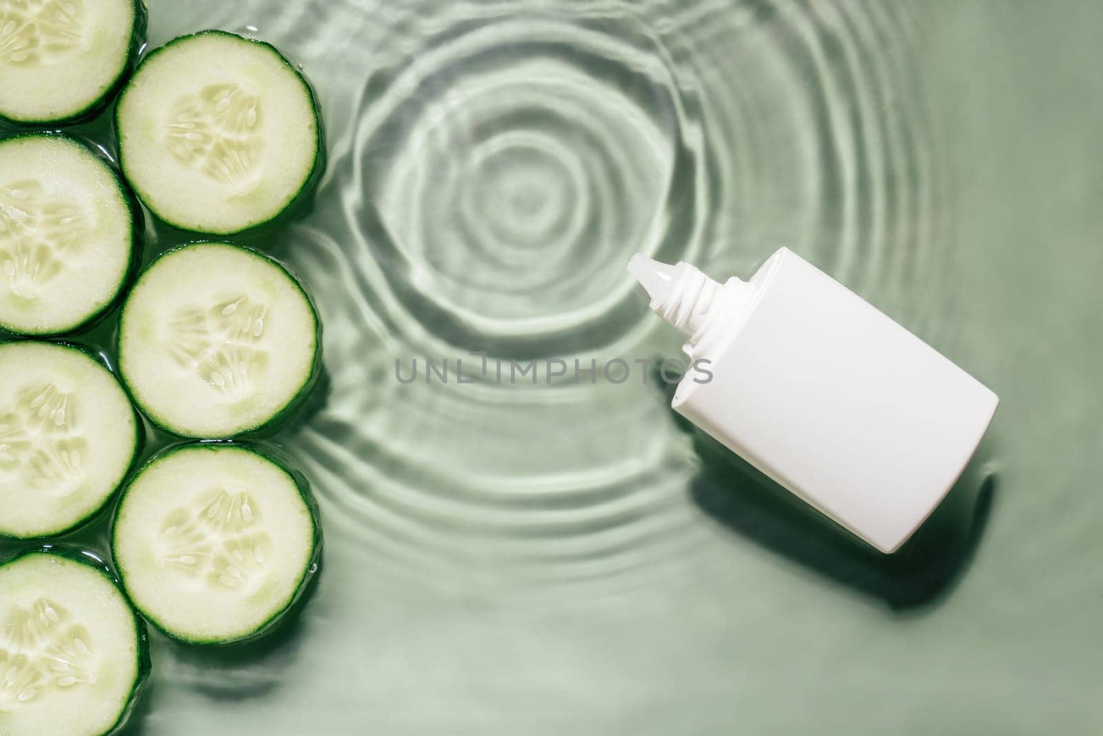 White beauty product cream vial tube bottle with rows of cucumber slices on surface of rippled transparent fresh green water gel with fleck, wave, shadow. Concentric expanding circles from fallen drop