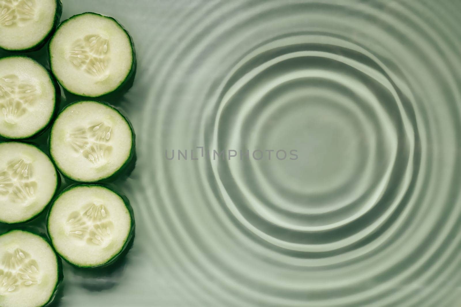 Cucumber slices lie on surface of rippled transparent fresh green exhilarant water gel with flecks, waves, shadow. Concentric expanding circles on surface from fallen drop. Health care. Copy space