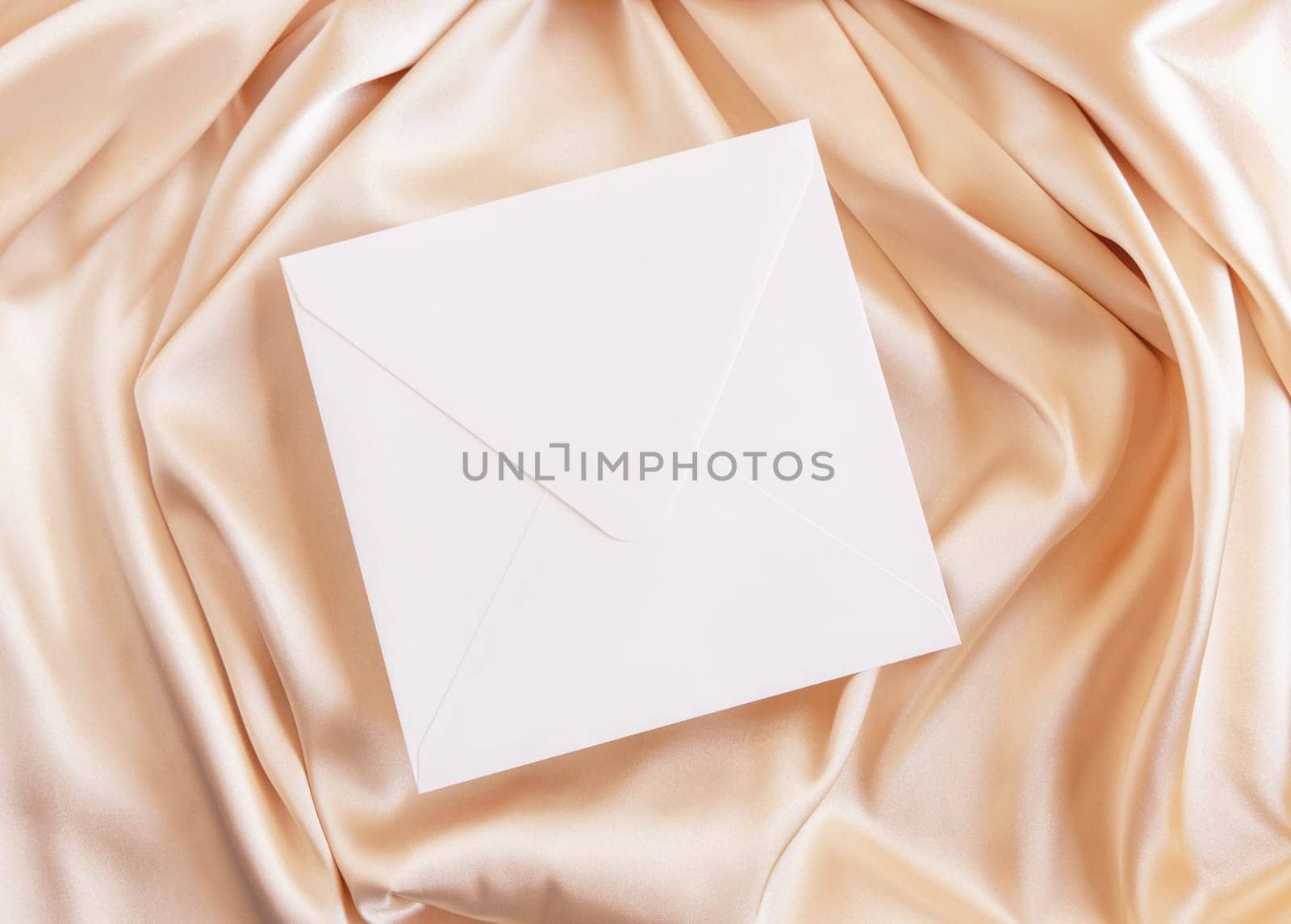 Square envelope on beige silky fabric drapery top view, mockup. Romantic scene with blank white envelope, greeting or wedding Invitation. .
