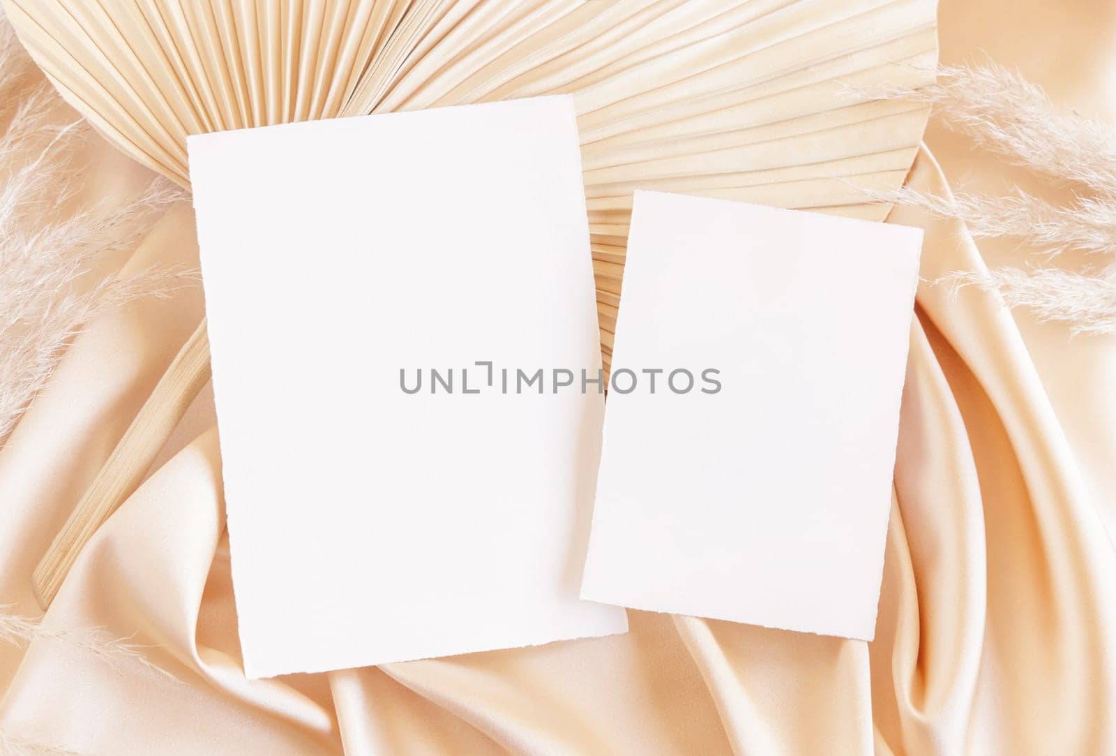 Blank cards on beige satin fabric and dried palm leaf close up, mockup. Boho scene with vertical paper cards. Bohemian or close to nature greeting or wedding Invitation. Romantic composition.