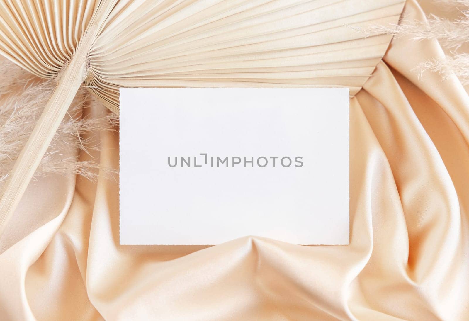 Blank card on beige satin fabric and dried palm leaf close up, mockup. Boho scene with horizontal paper card. Bohemian or close to nature greeting or wedding Invitation. Romantic composition.