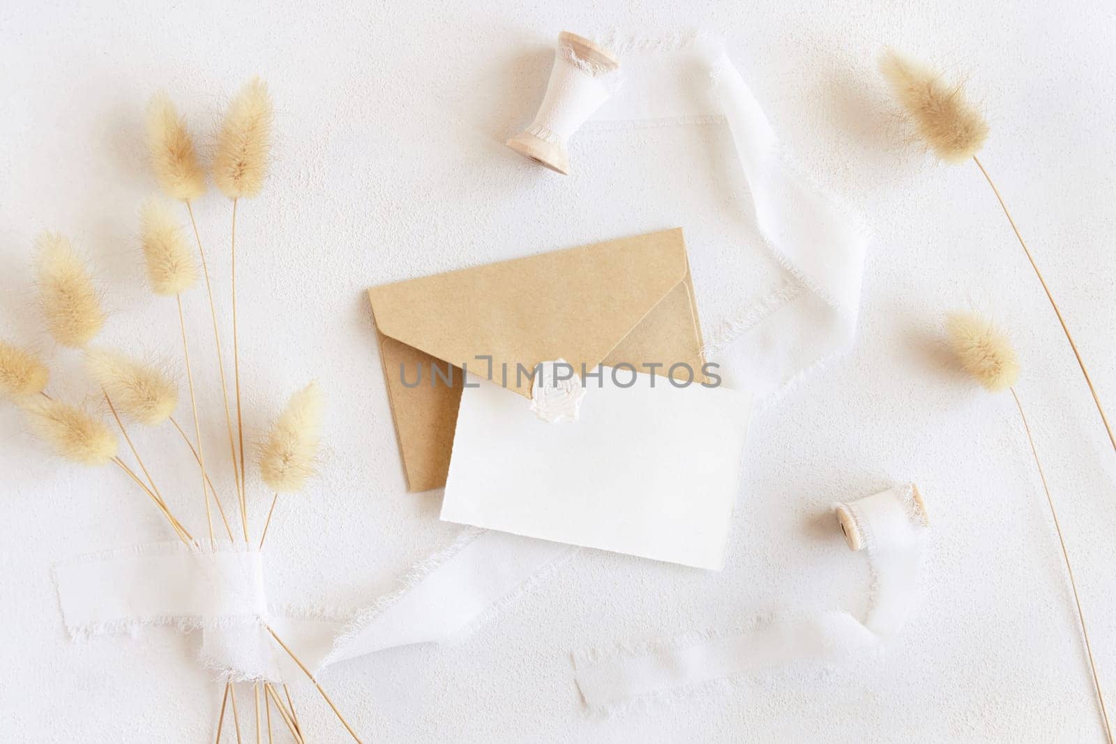 Blank card and envelope near silk ribbons and dried hare's tail grass top view on white, mockup. Boho romantic scene with horizontal cards. Bohemian or close to nature greeting or wedding Invitation