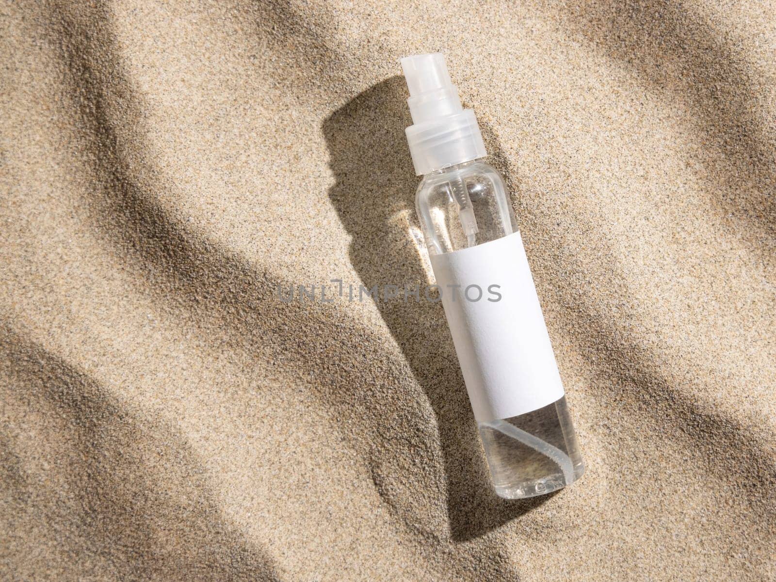 Spray bottle with blank label on beige sand top view, hard light. Package mockup. Natural skincare or SPA product. Facial cosmetics for beauty routine. Zen style with waves on sand, minimal aesthetic