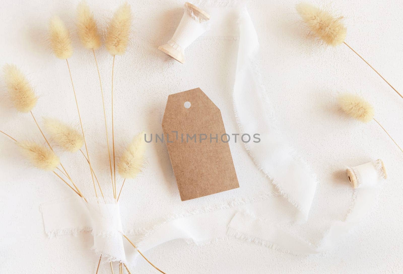 Blank gift tag near silk ribbons and dried hare's tail grass top view on white, mockup. Boho romantic scene with kraft paper gift tag. Bohemian or close to nature greeting
