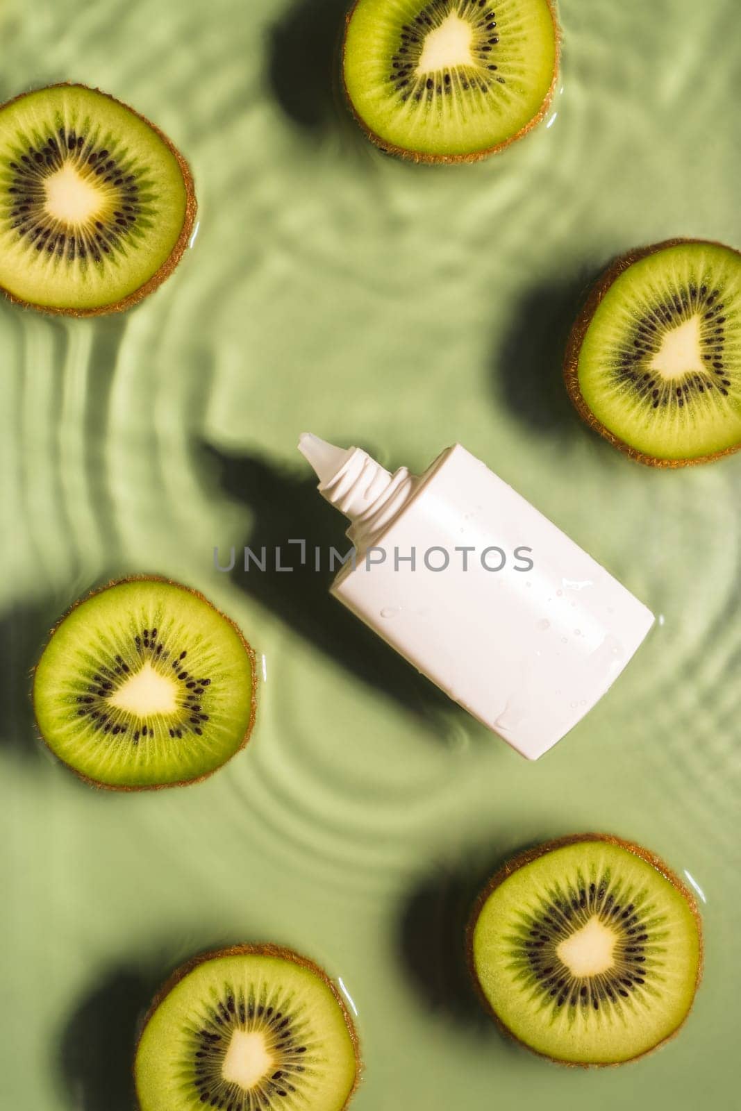 White beauty product sunscreen cream vial tube bottle in transparent fresh exhilarant wave water with flecks, fruit tropical kiwi slices. Body skin care protection in summer beach concept. Vertical