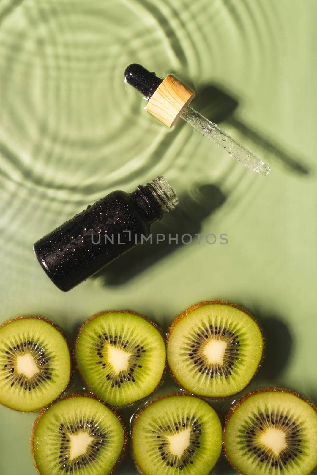 Facial pipette serum black bottle, whey beauty product near fruit tropical kiwi slices in water splashing fresh transparent, flecks. Top view. Organic skin face and body moisturizing. Vertical