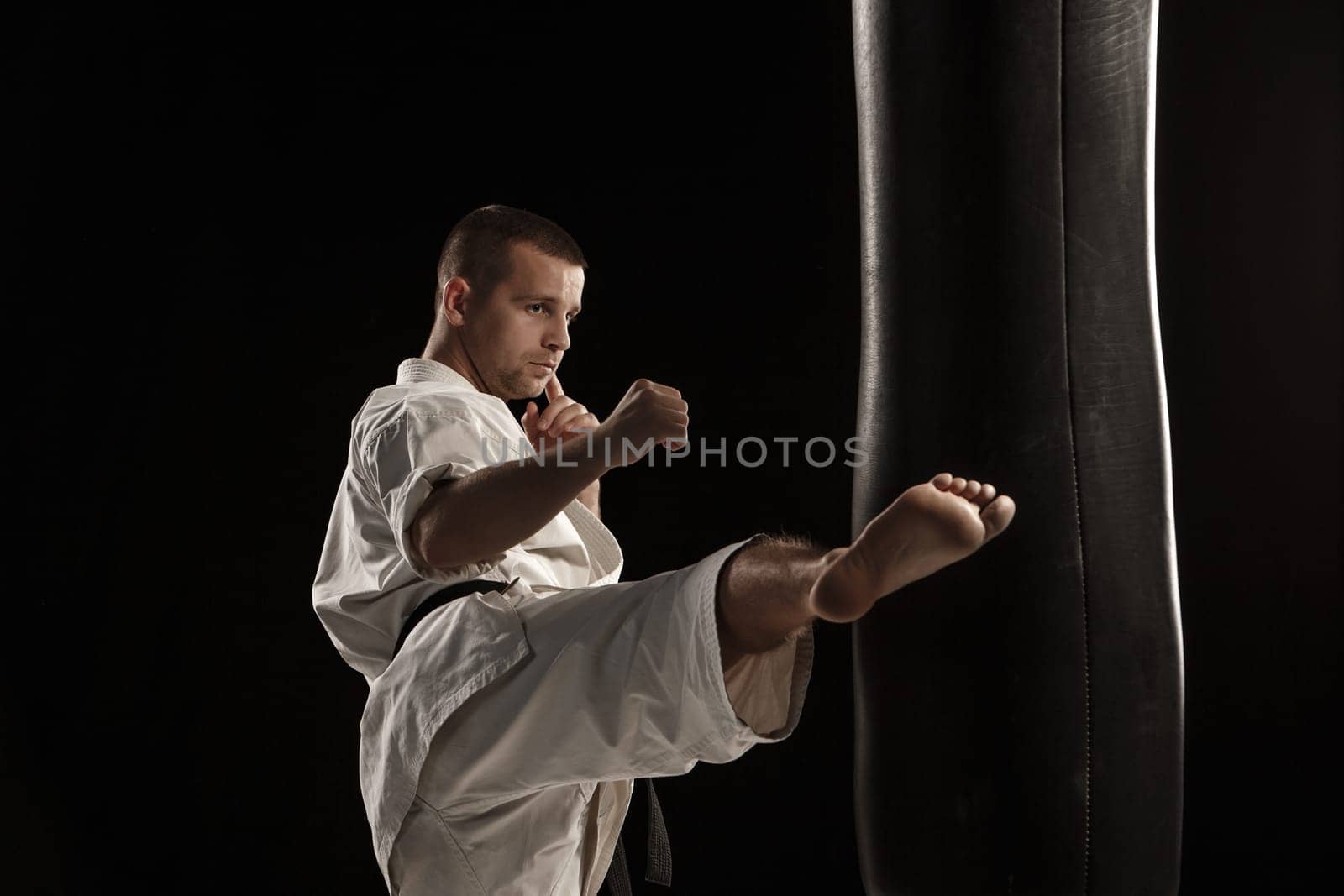 Man in white kimono training karate round kick in a punching bag over black background