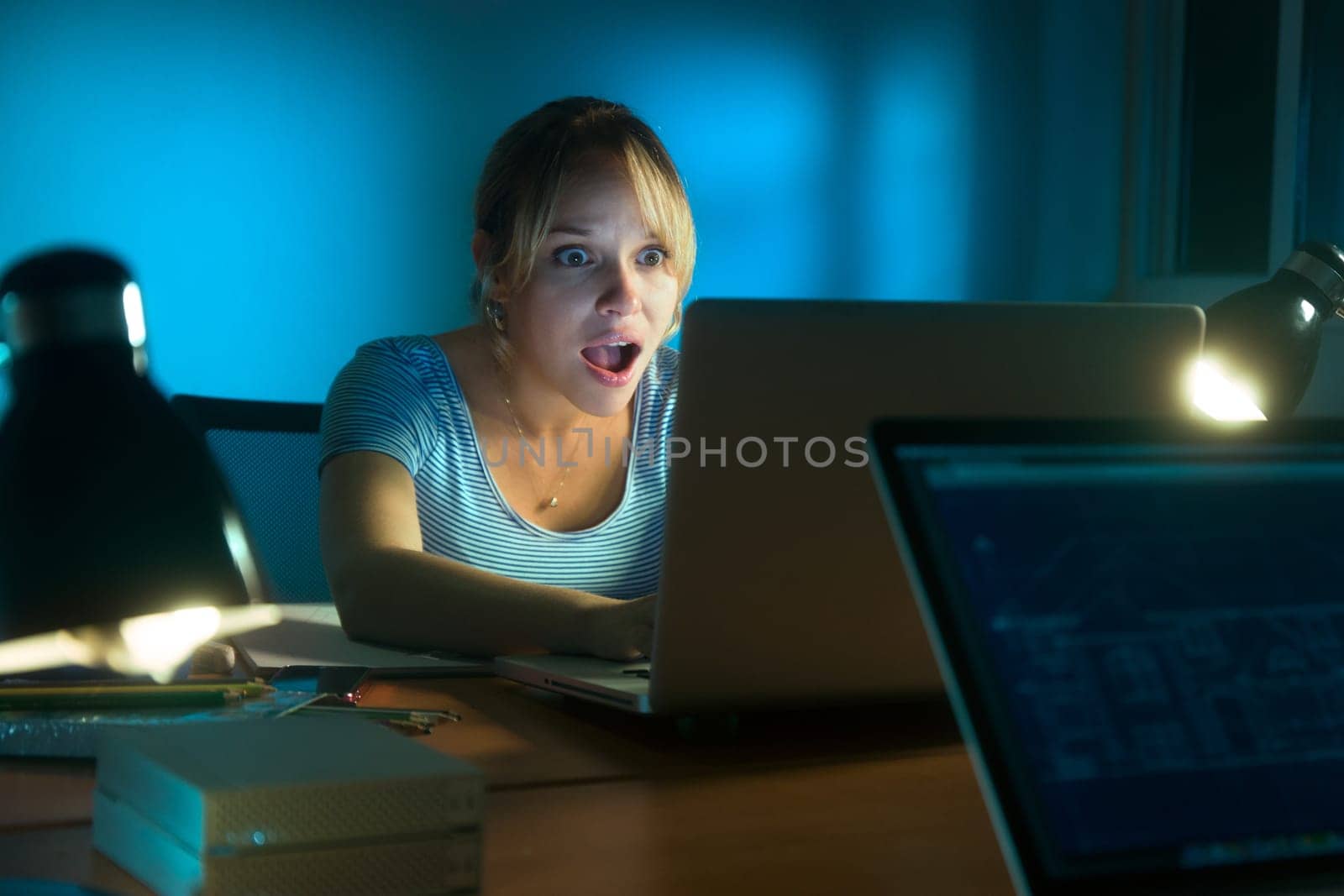Beautiful woman working late at night in office, surfing the web and reading a post on social network with laptop computer. The girl looks at the screen with open mouth