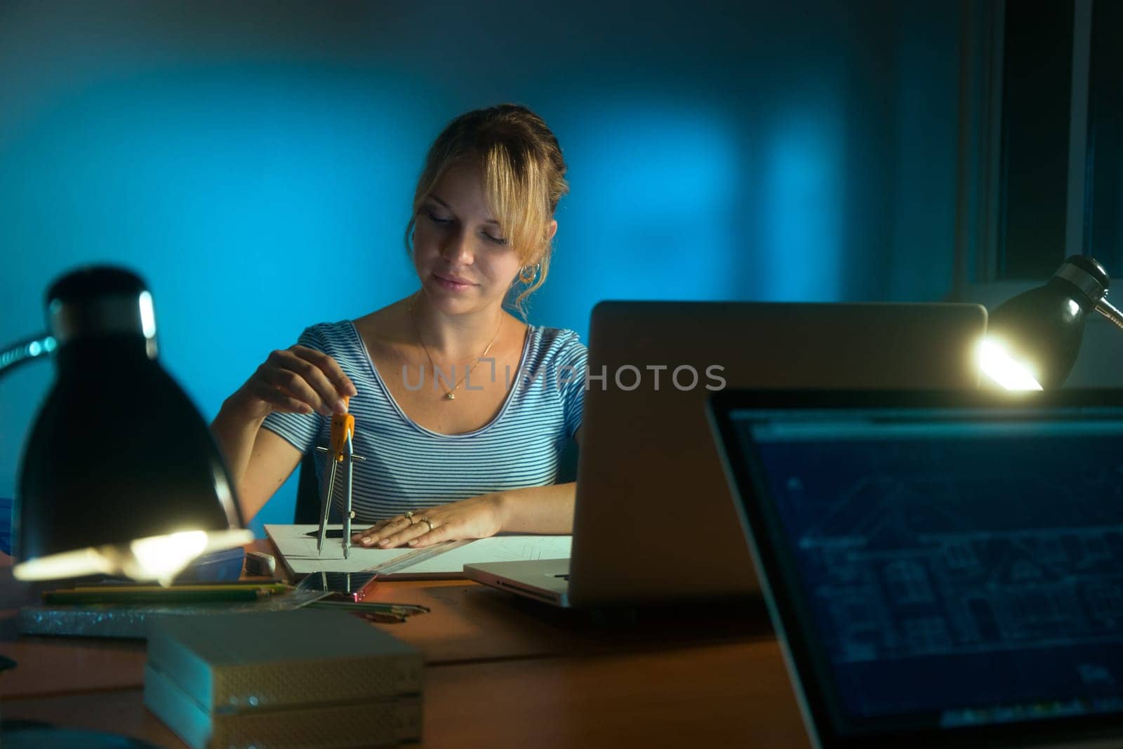 Beautiful woman working as interior designer, staying late at night in office with drawings and laptop computer to complete a project. The girl draws a circle with compasses