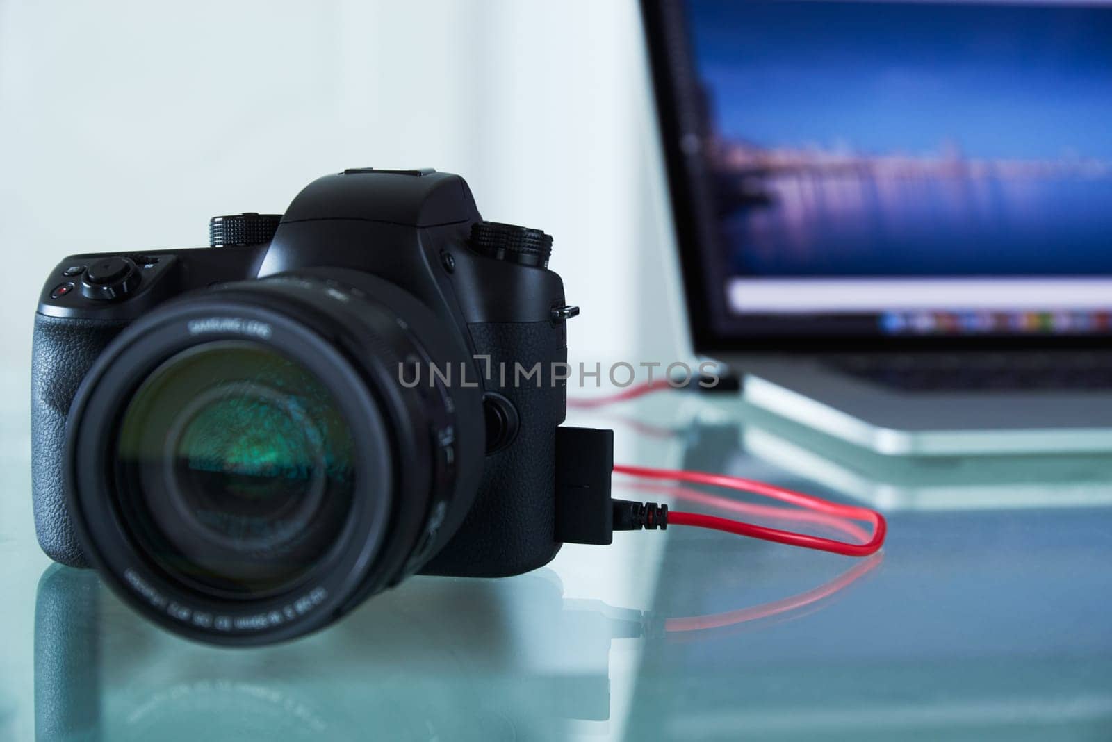 Still life of dslr camera connected with USB cable to laptop computer on glass table. The photo camera is transferring images to PC in background. Copy space
