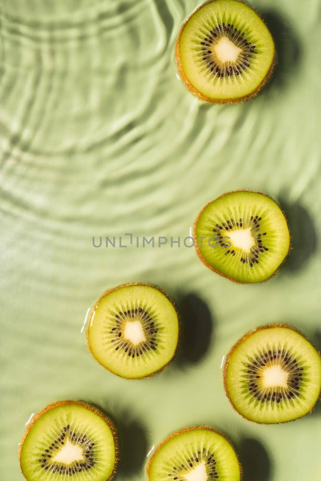 Kiwi slices lie on surface of rippled transparent fresh green exhilarant water gel with flecks, waves, shadow. Concentric expanding circles on surface from fallen drop. Vertical. Copy space