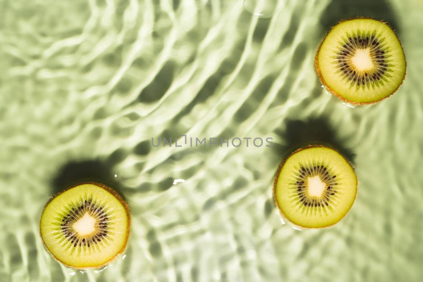 Kiwi slices lie on surface of rippled transparent fresh green exhilarant water gel with flecks, waves, shadow. Concentric expanding circles on surface from fallen drop. Health care. Copy space