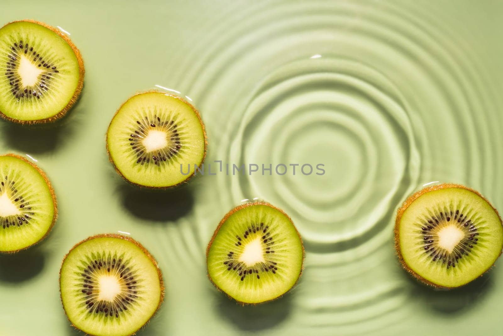 Kiwi slices lie on surface of rippled transparent fresh green exhilarant water gel with flecks, waves, shadow. Concentric expanding circles on surface from fallen drop. Health care. Copy space