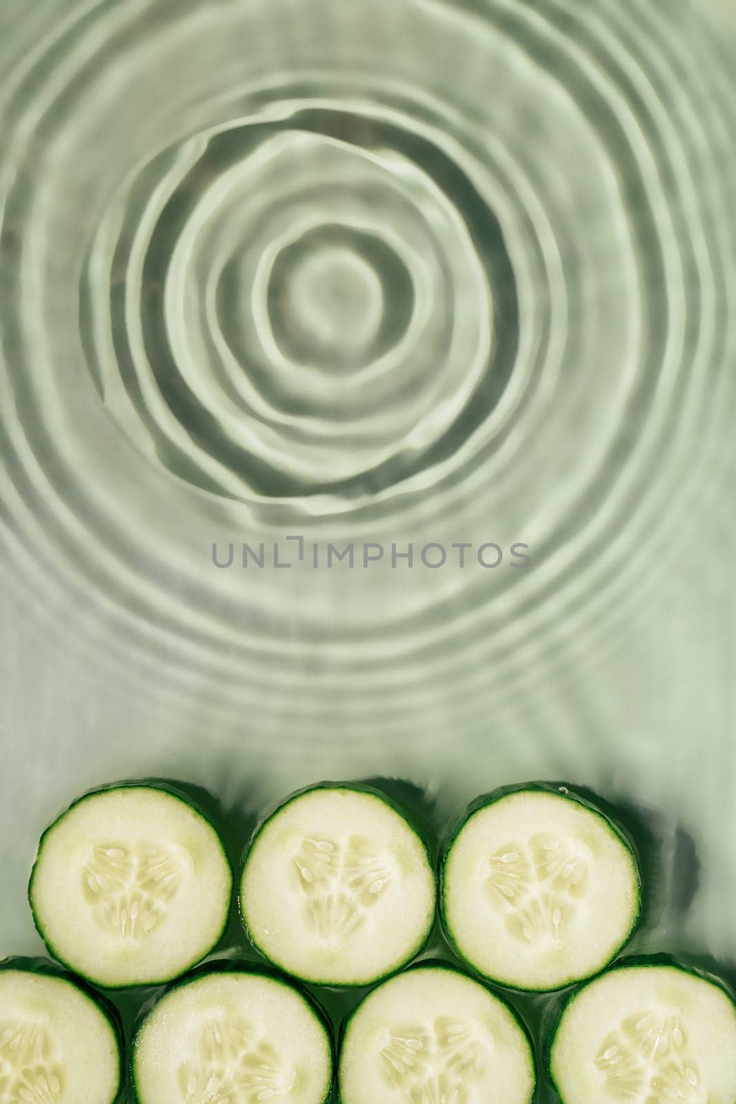 Cucumber slices lie on surface of rippled transparent fresh green exhilarant water gel with flecks, waves, shadow. Expanding circles on surface from fallen drop. Health care. Copy space. Vertical
