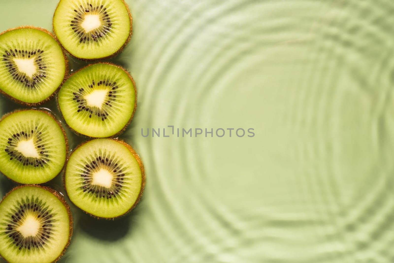 Kiwi slices lie on surface of rippled transparent fresh green exhilarant water gel with flecks, waves, shadow. Concentric expanding circles on surface from fallen drop. Health care. Copy space